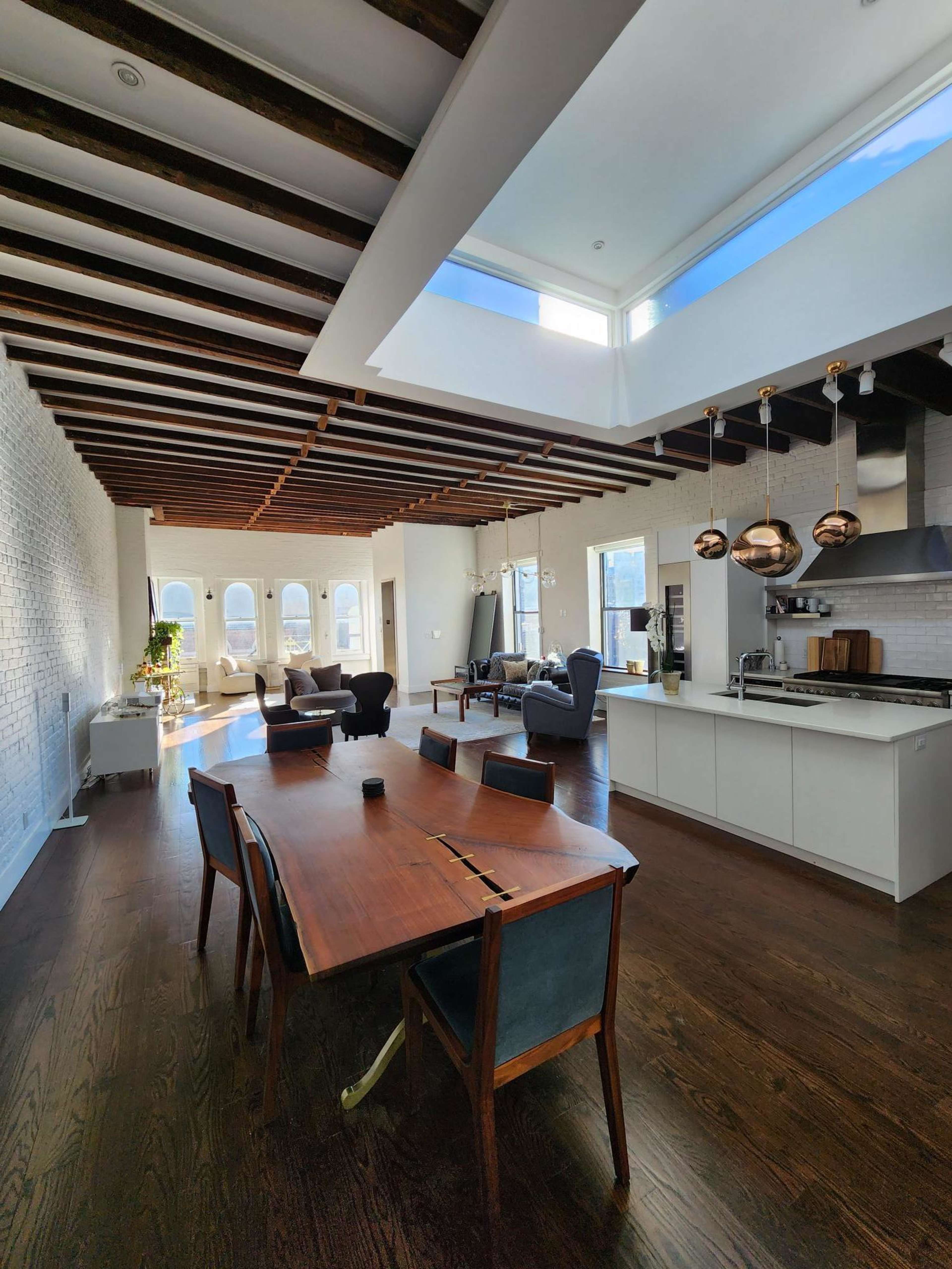 A spacious, modern living area features a large dining table, sleek kitchen, and high ceilings with exposed beams and large windows.