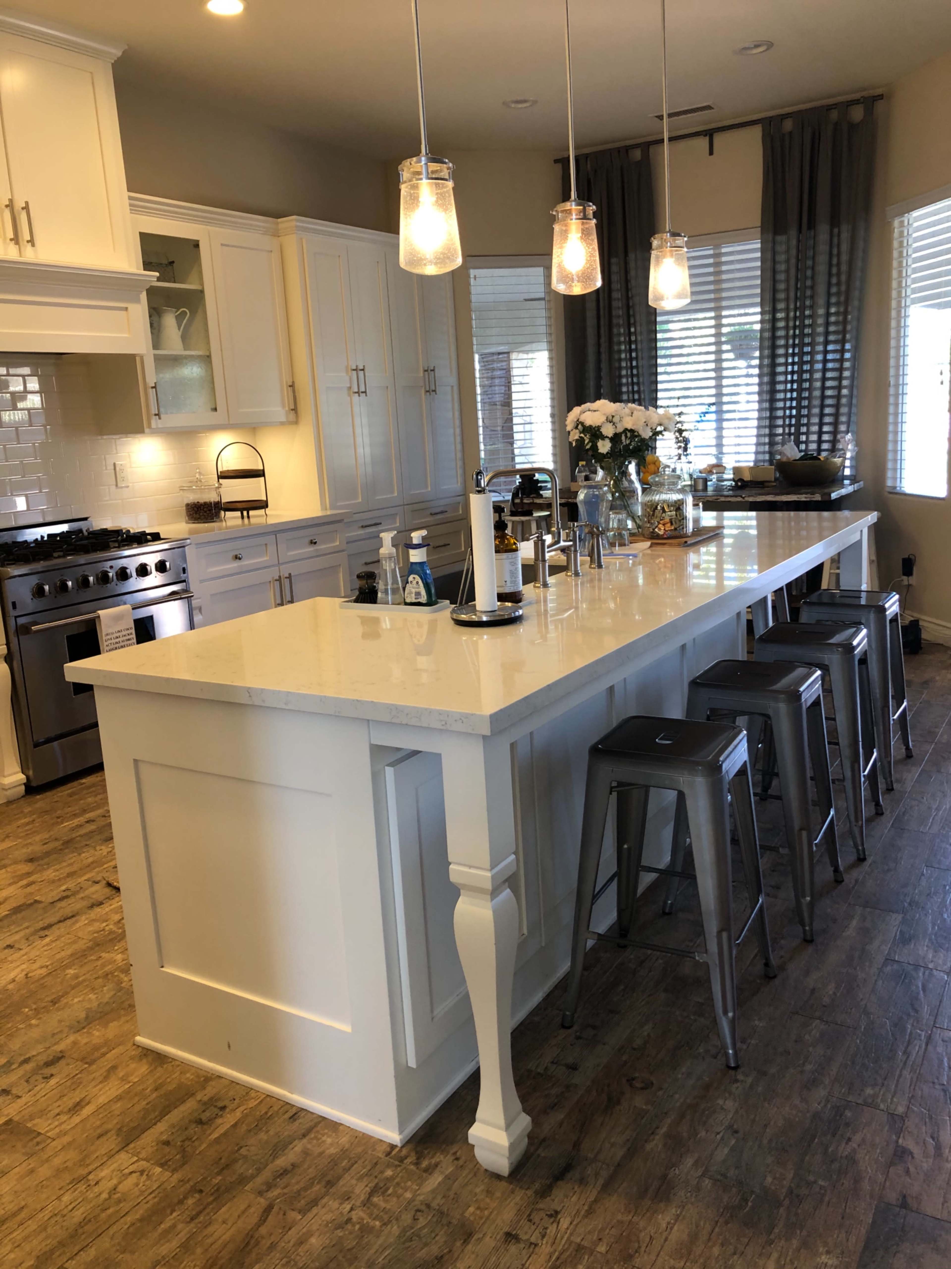A modern kitchen with a spacious island, bar stools, and stainless steel appliances, featuring white cabinetry and a light countertop.