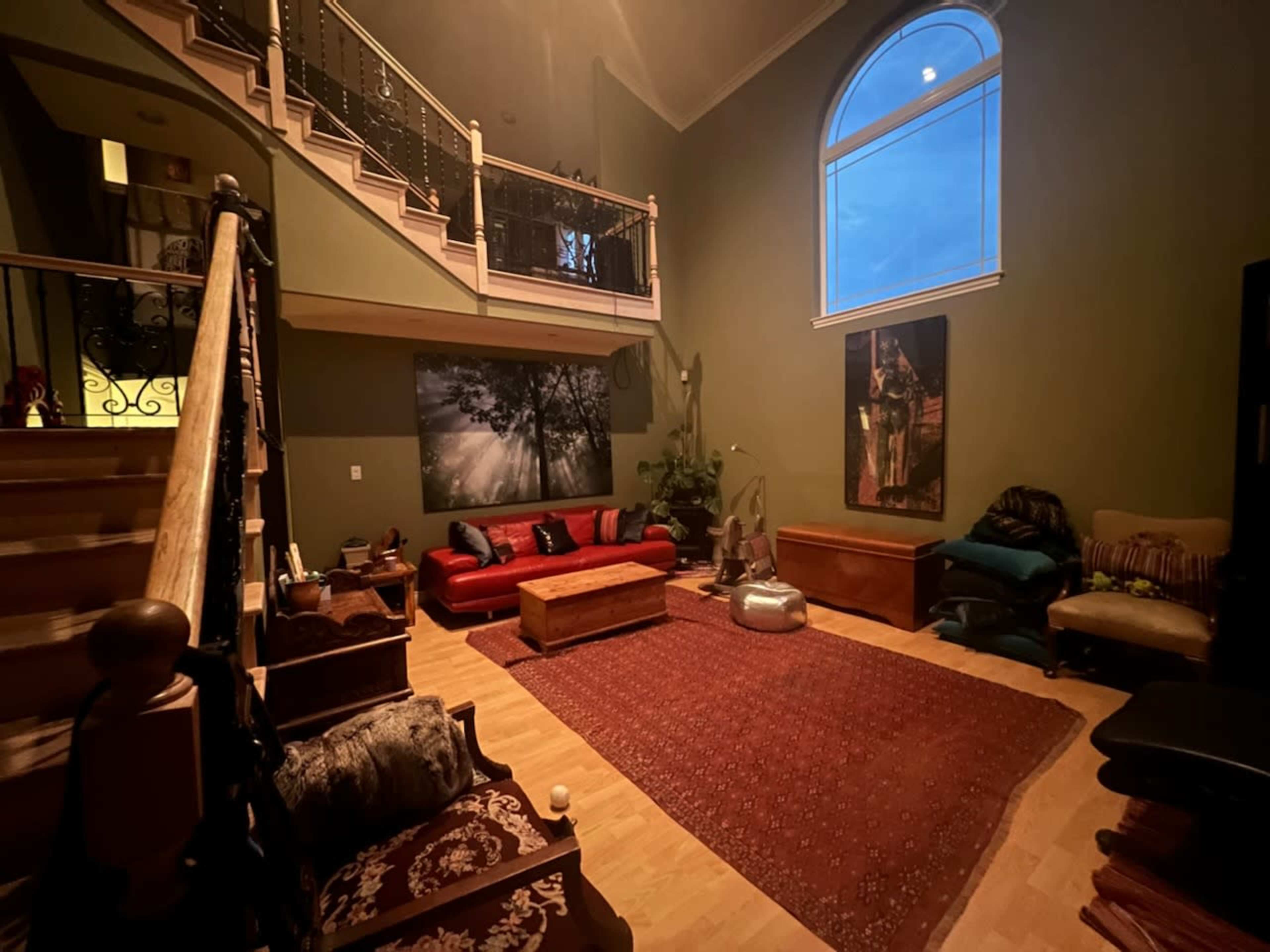 The image shows a spacious living room with dark green walls, a large window letting in twilight, a red sofa set, a wooden coffee table, and various decorative items scattered around the floor.