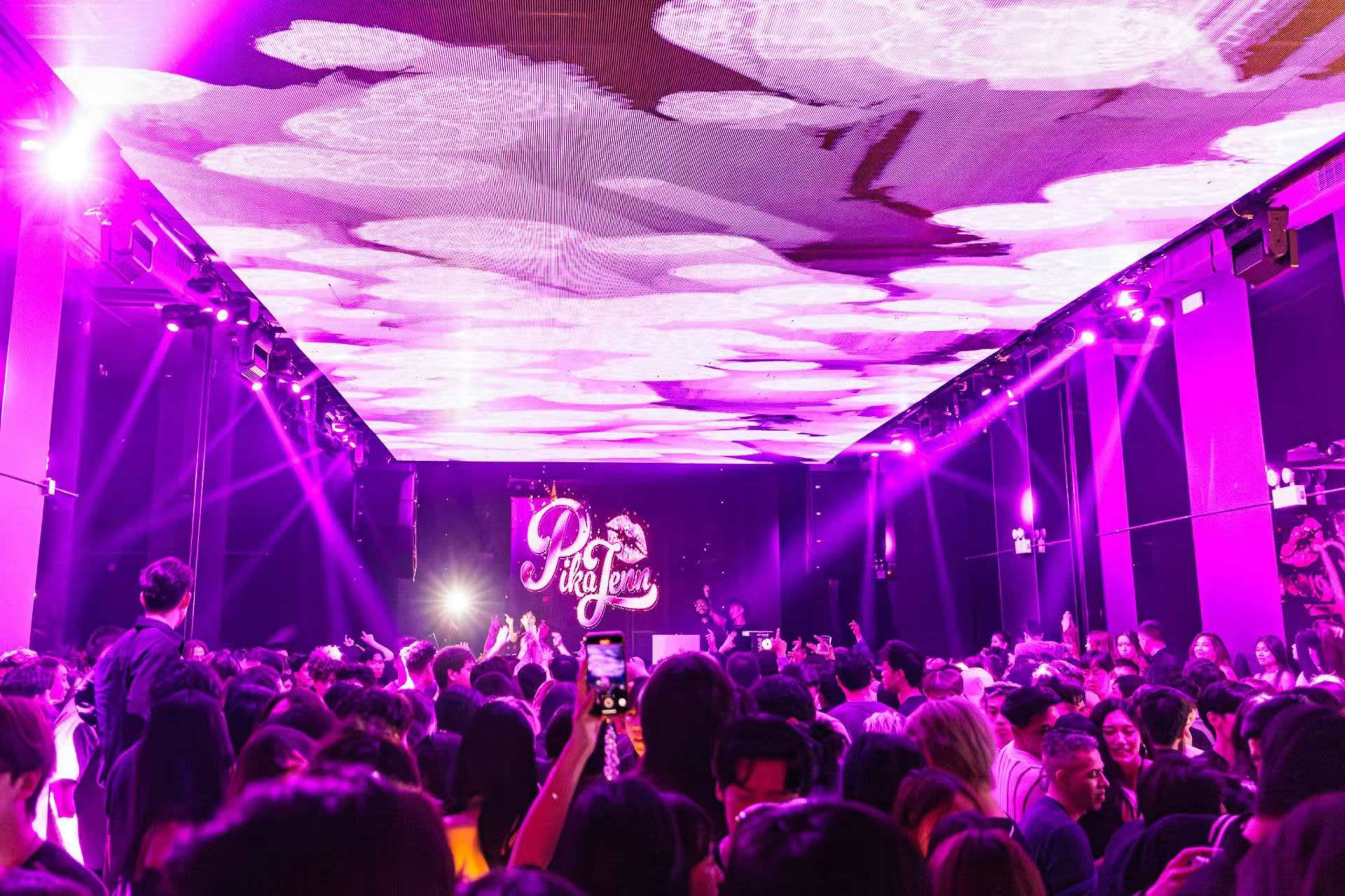 A crowded nightclub scene features a vibrant light display overhead with people dancing and using their phones to capture the moment.