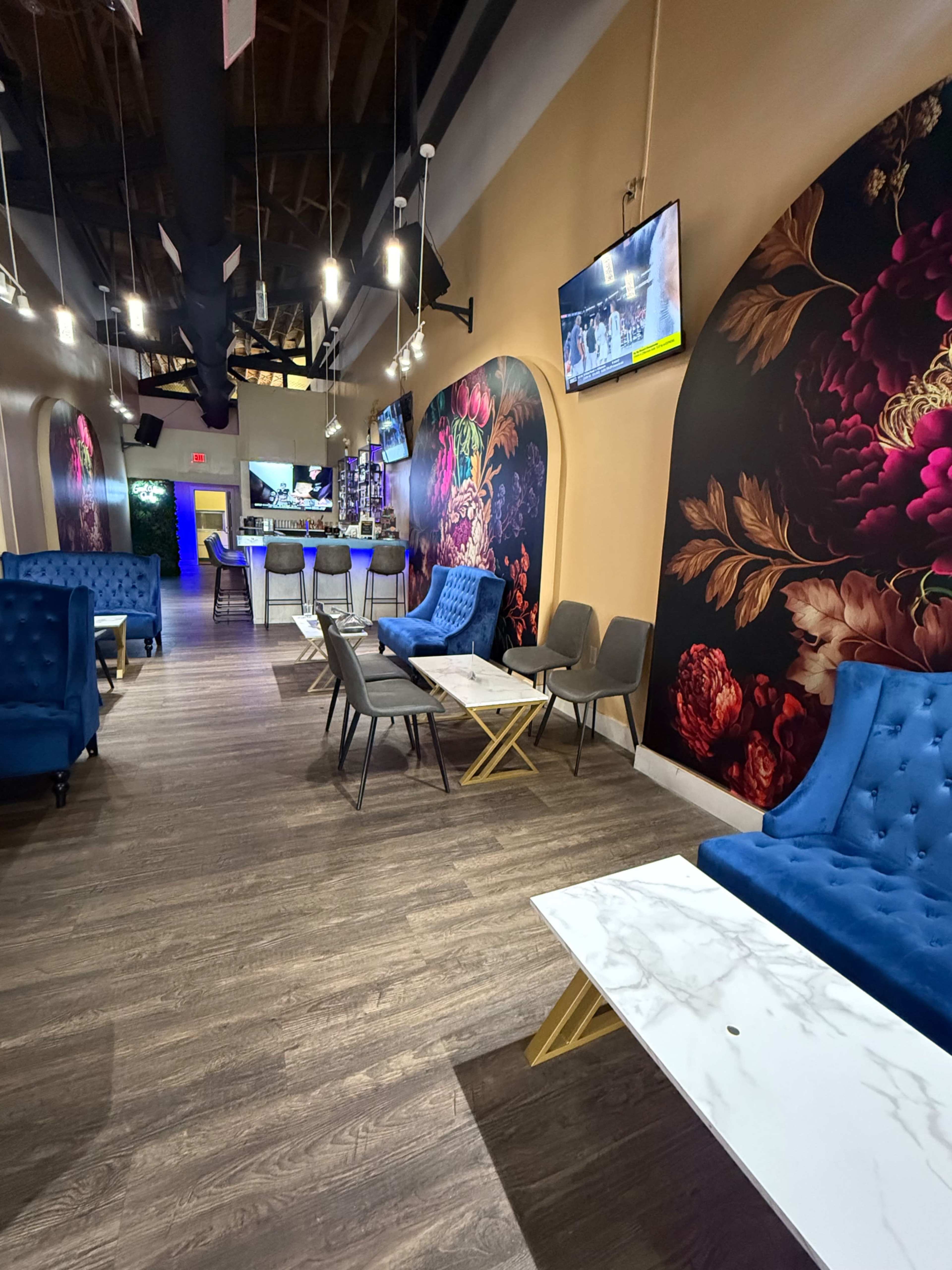 The image shows a brightly lit lounge with floral wallpaper, plush blue seating, and several tables arranged along a wooden floor.