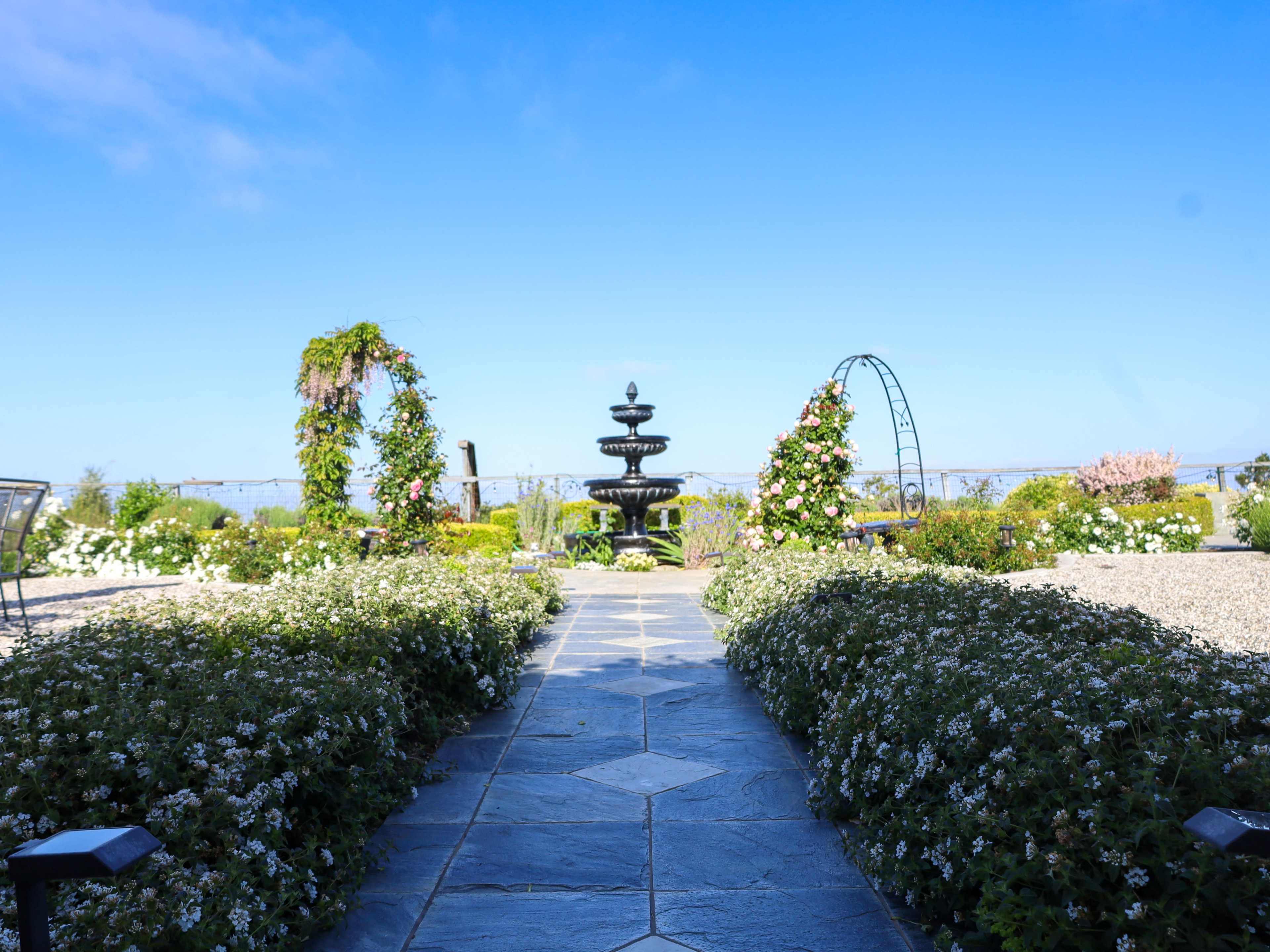 A stone pathway leads through flowerbeds to a central fountain surrounded by arches adorned with greenery under a clear blue sky.