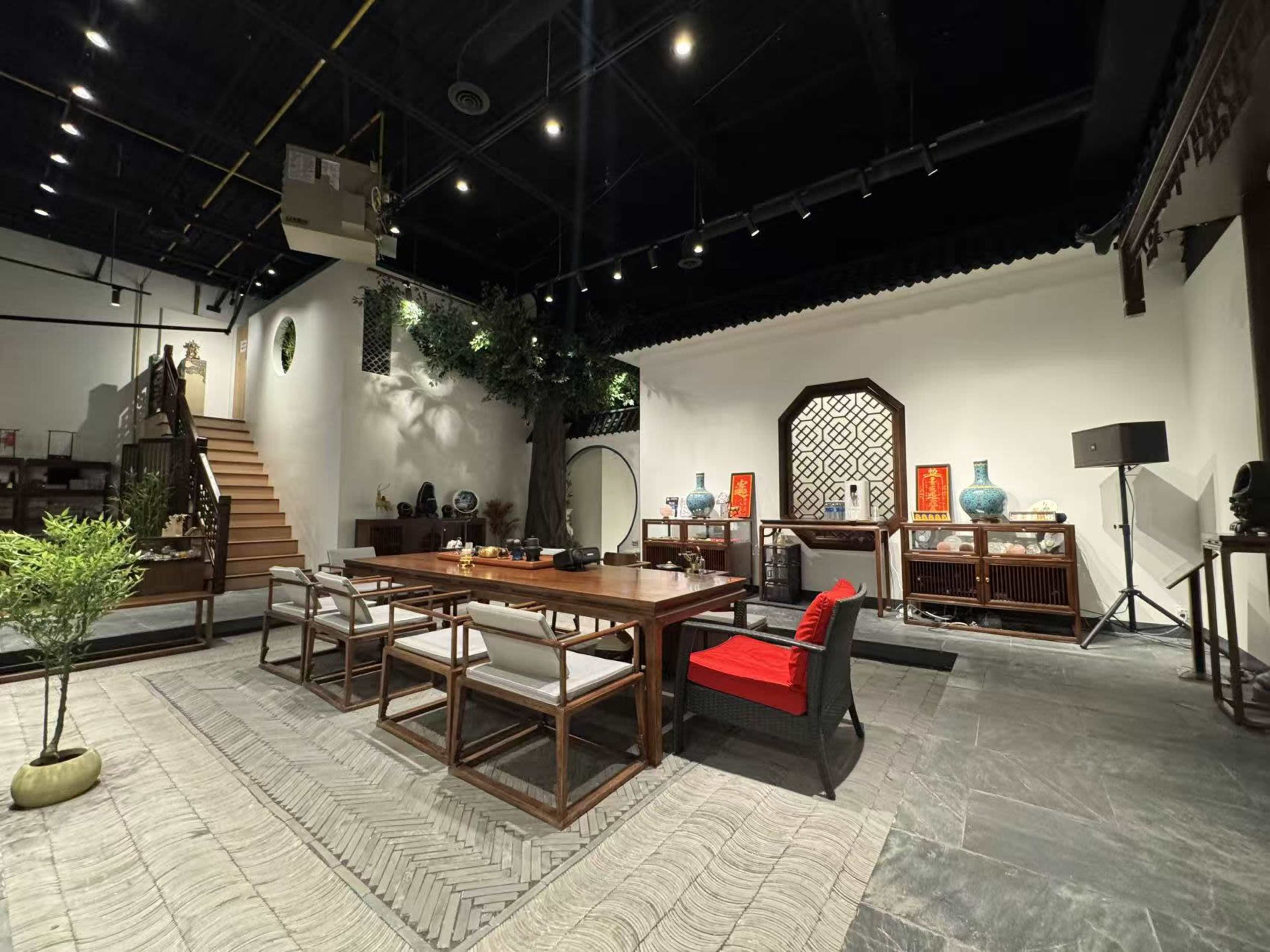 The image shows a spacious modern room with a large wooden dining table surrounded by chairs, decorated with plants and traditional furnishings, and featuring a staircase and decorative elements on the walls.