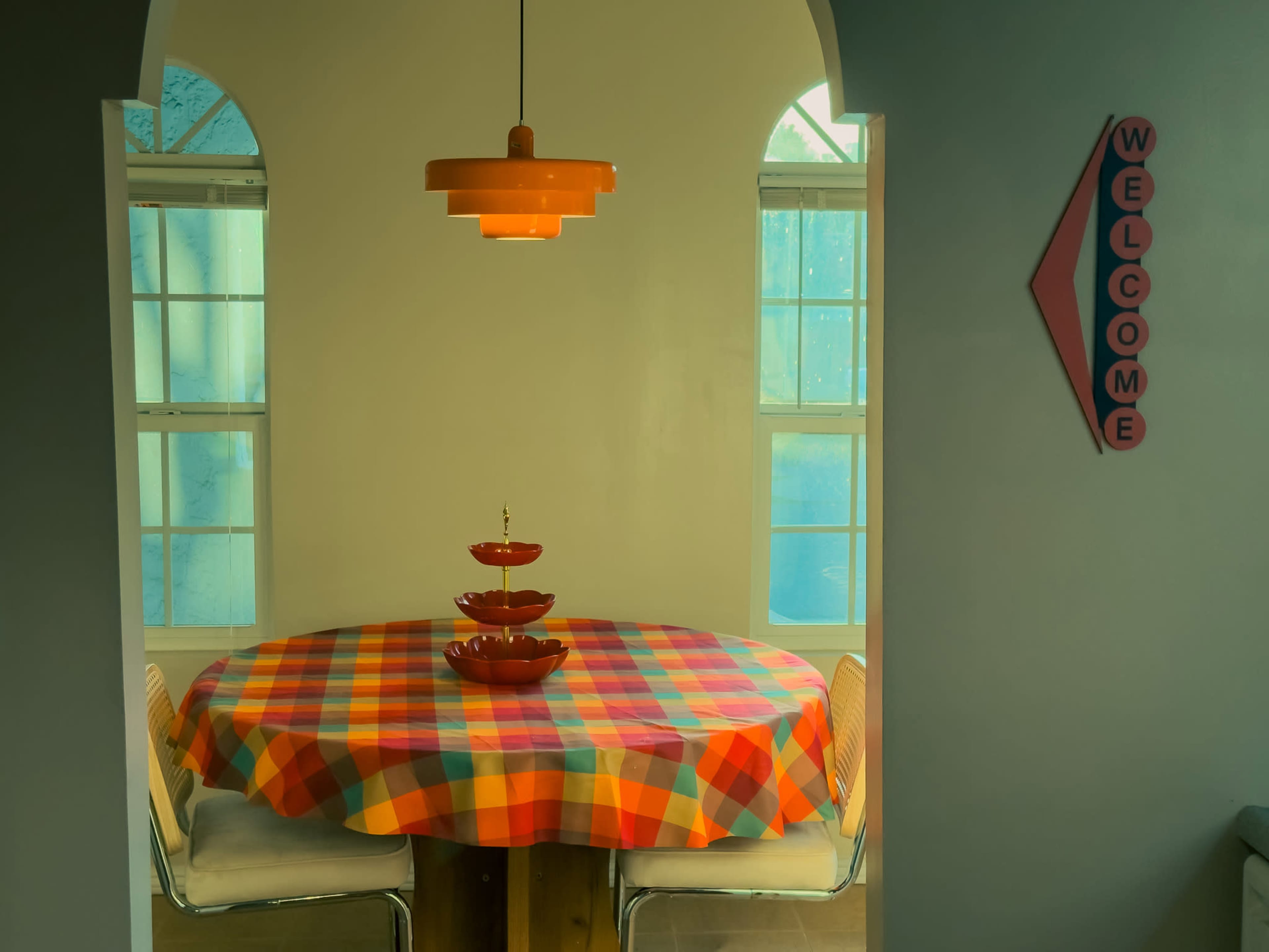 A round table covered with a colorful checkered tablecloth is set in a well-lit room, with a decorative three-tiered serving tray and a warm pendant light overhead.