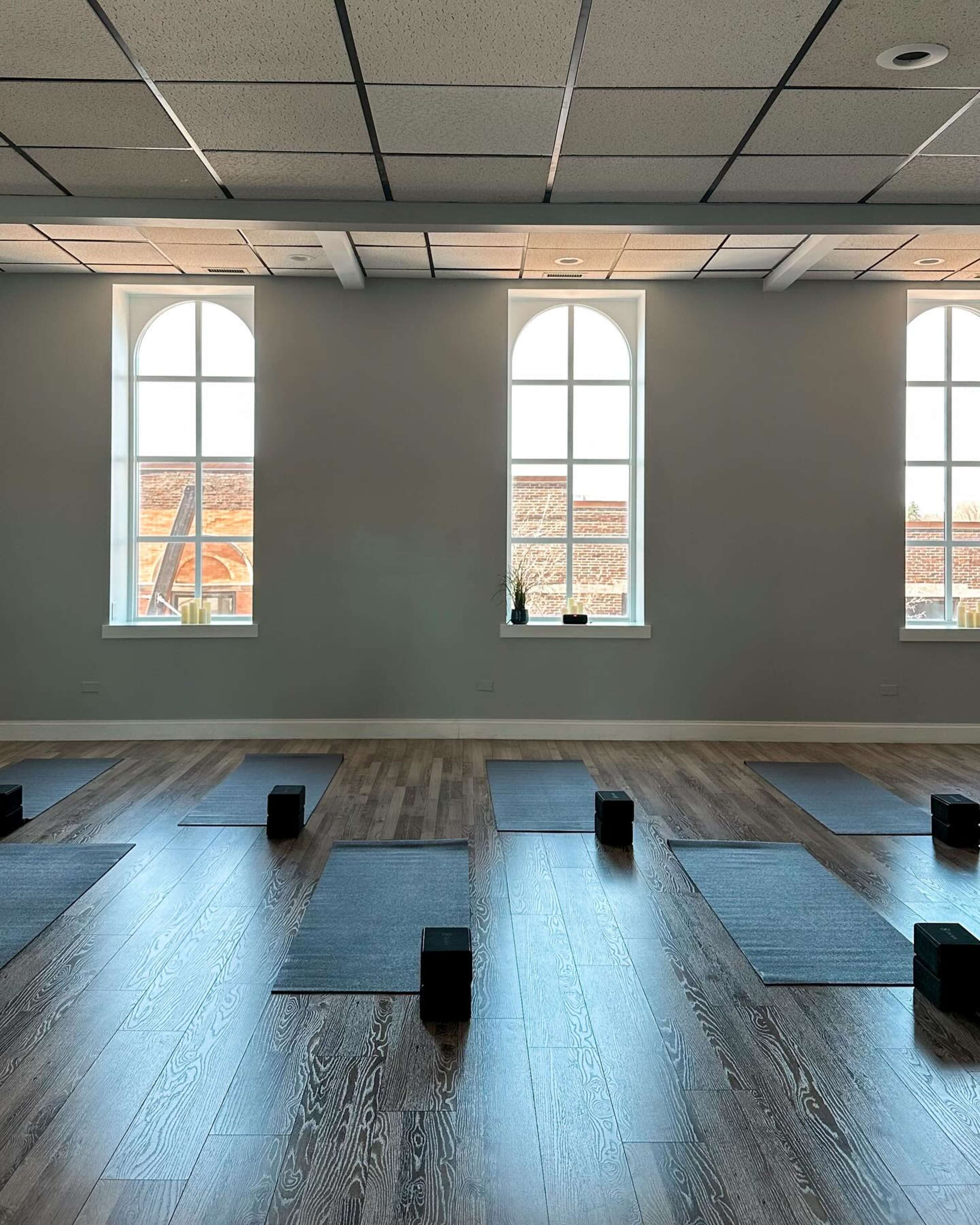 The image shows a spacious room with yoga mats and blocks arranged in parallel rows, illuminated by natural light from large windows.