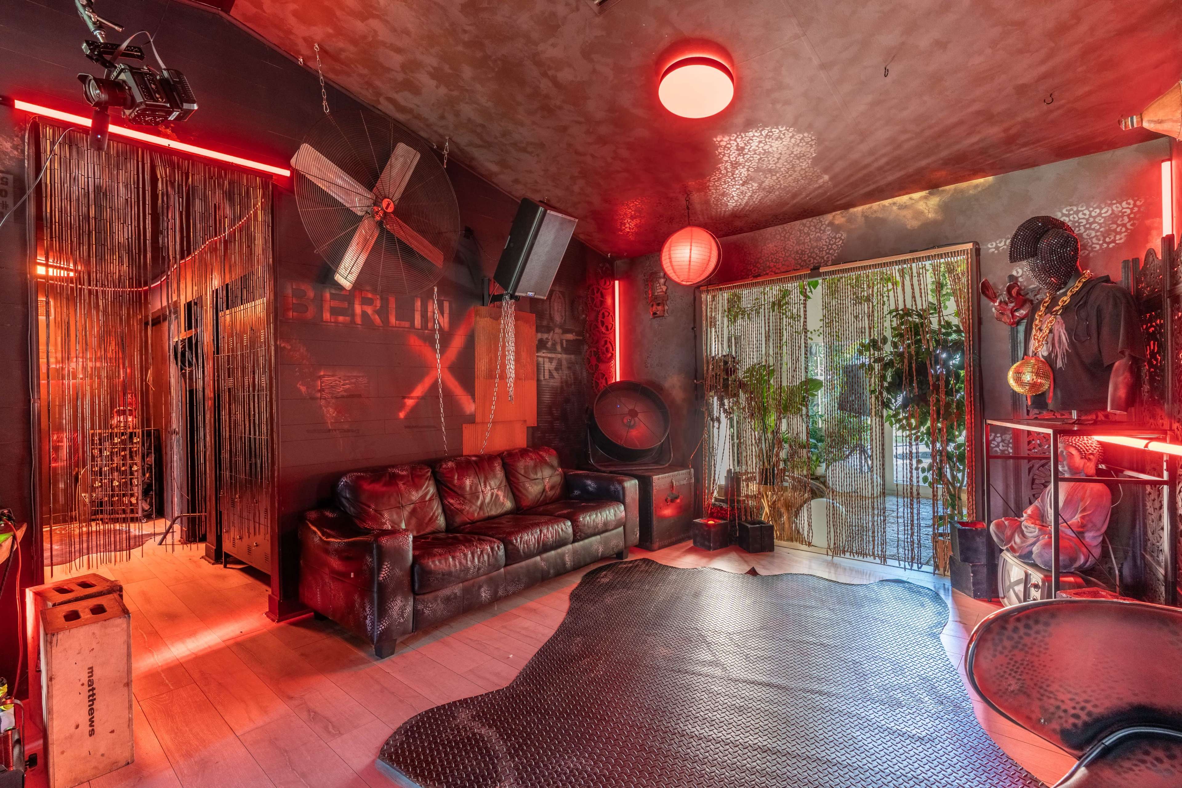The image shows a stylized room featuring a red-lit aesthetic, with a mix of industrial decor, a leather sofa, and plants visible through a doorway.
