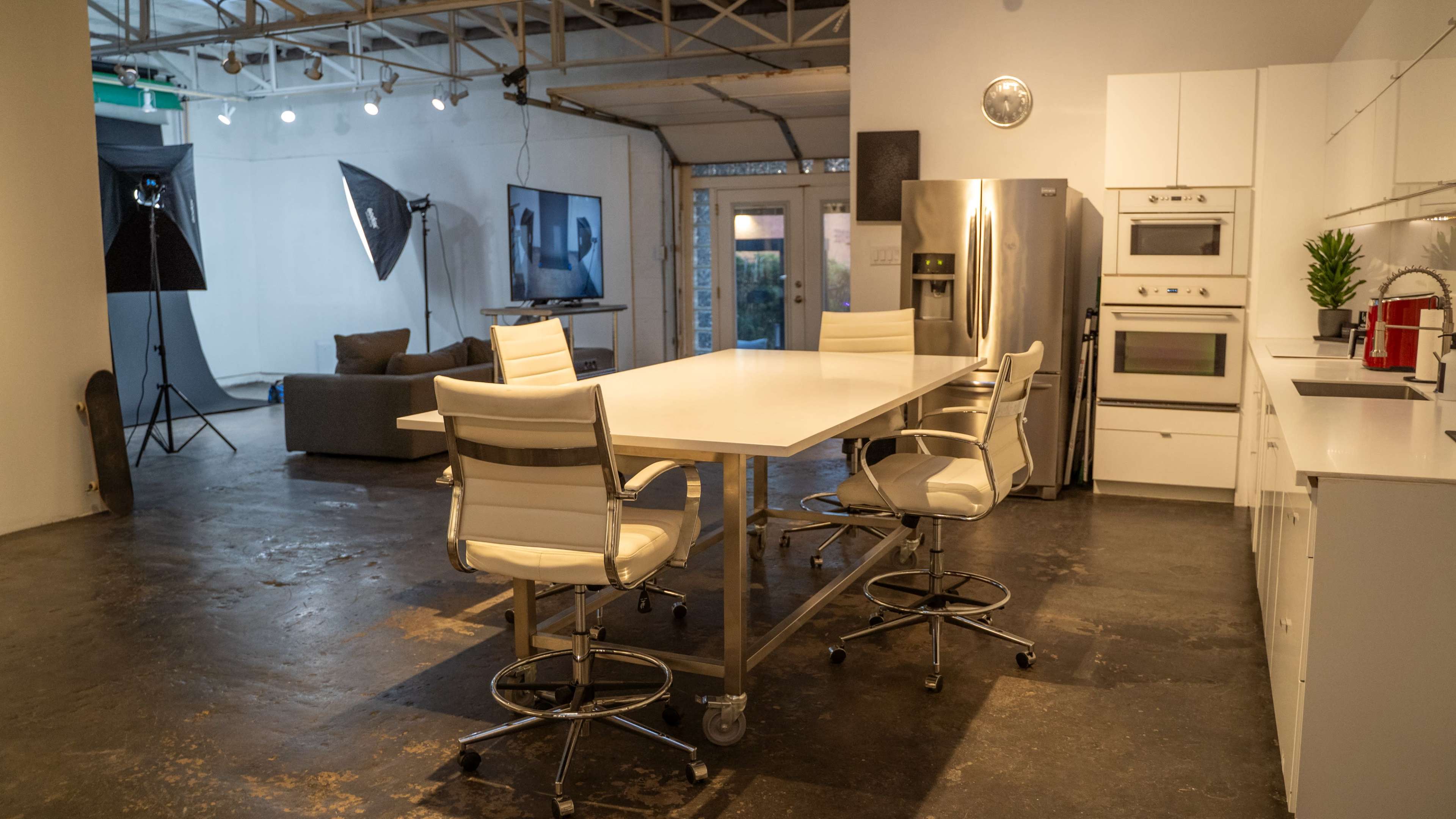 A modern kitchen with a large white table and four high-backed chairs, surrounded by stainless steel appliances and an open space featuring a couch and a camera setup.