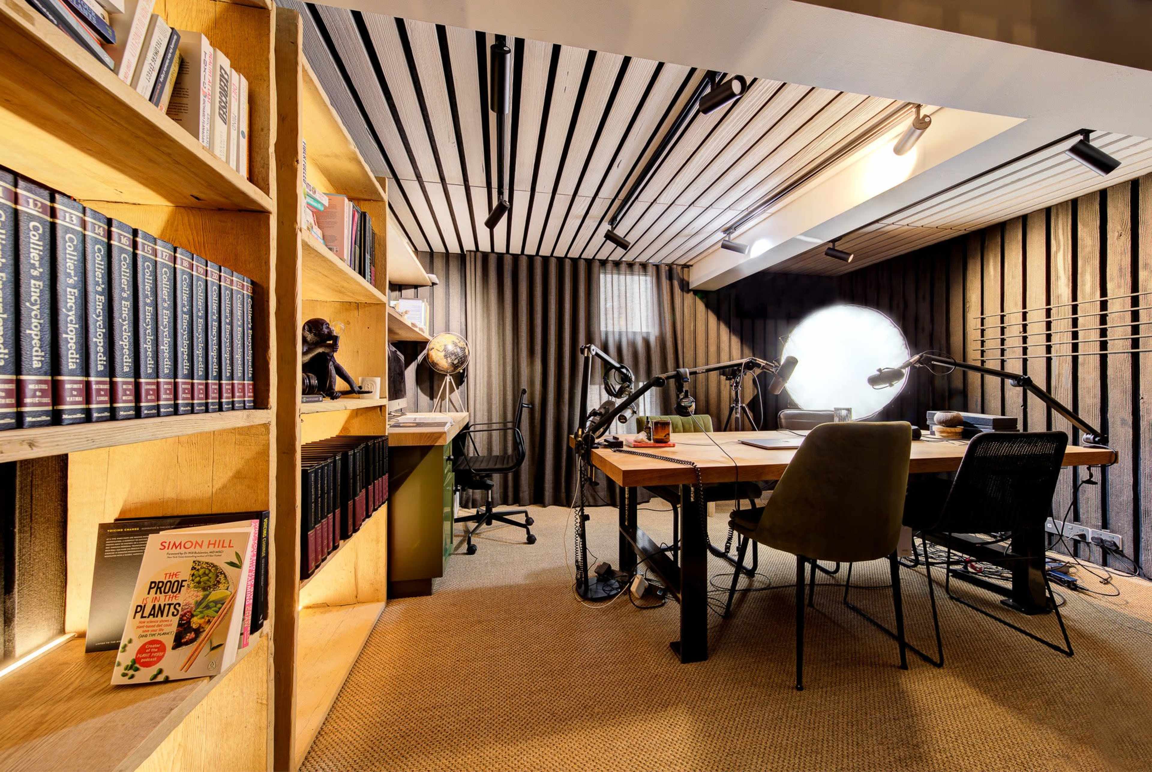 The image shows a modern office space featuring a wooden bookshelf filled with books, a large desk with two chairs, and various desk lamps under striped ceiling panels.