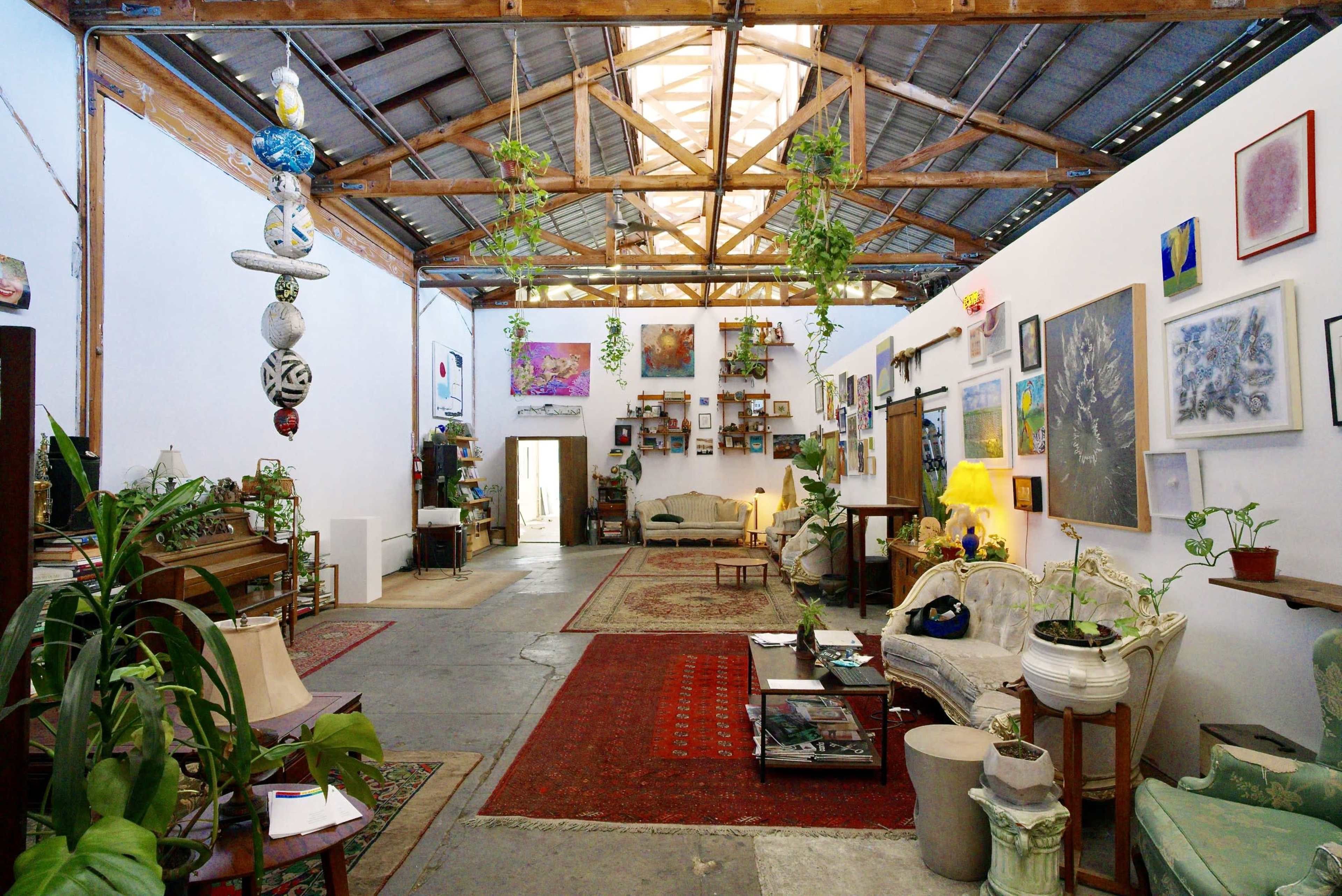 The image shows an open gallery space featuring a mix of artwork and plants, with furniture arranged for seating along the walls under a high ceiling.