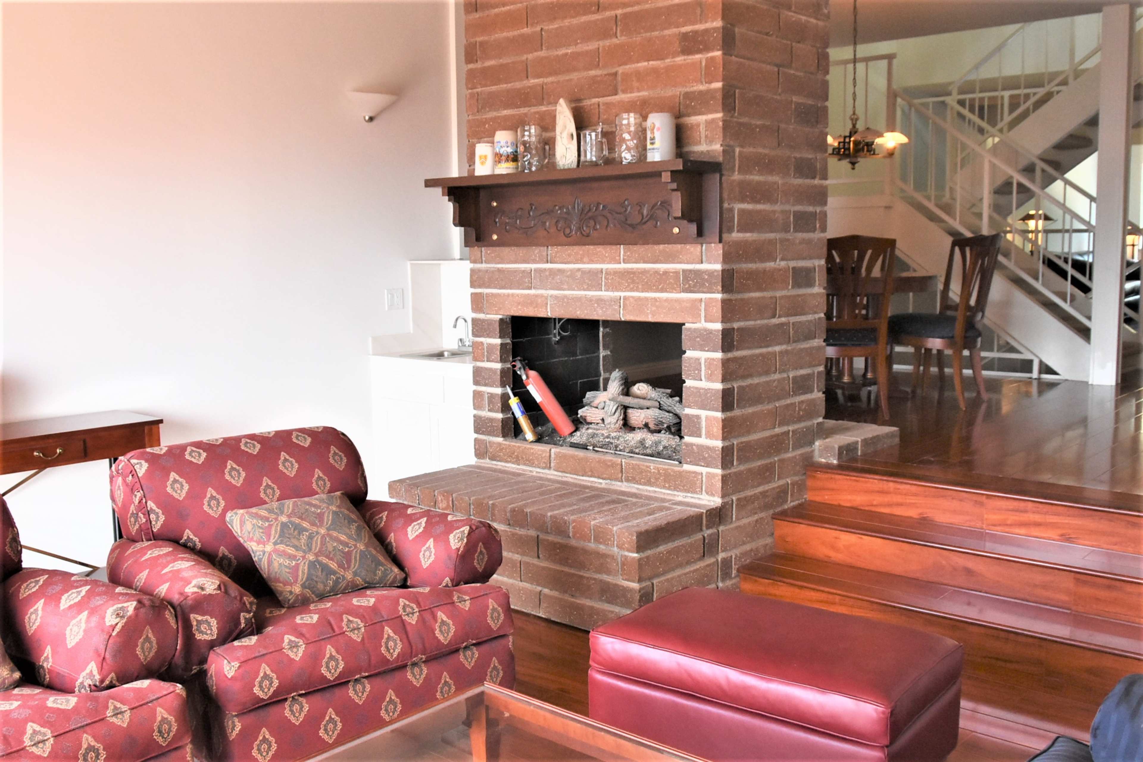 The image shows a living room with a brick fireplace surrounded by a red patterned sofa and a wooden dining area in the background.