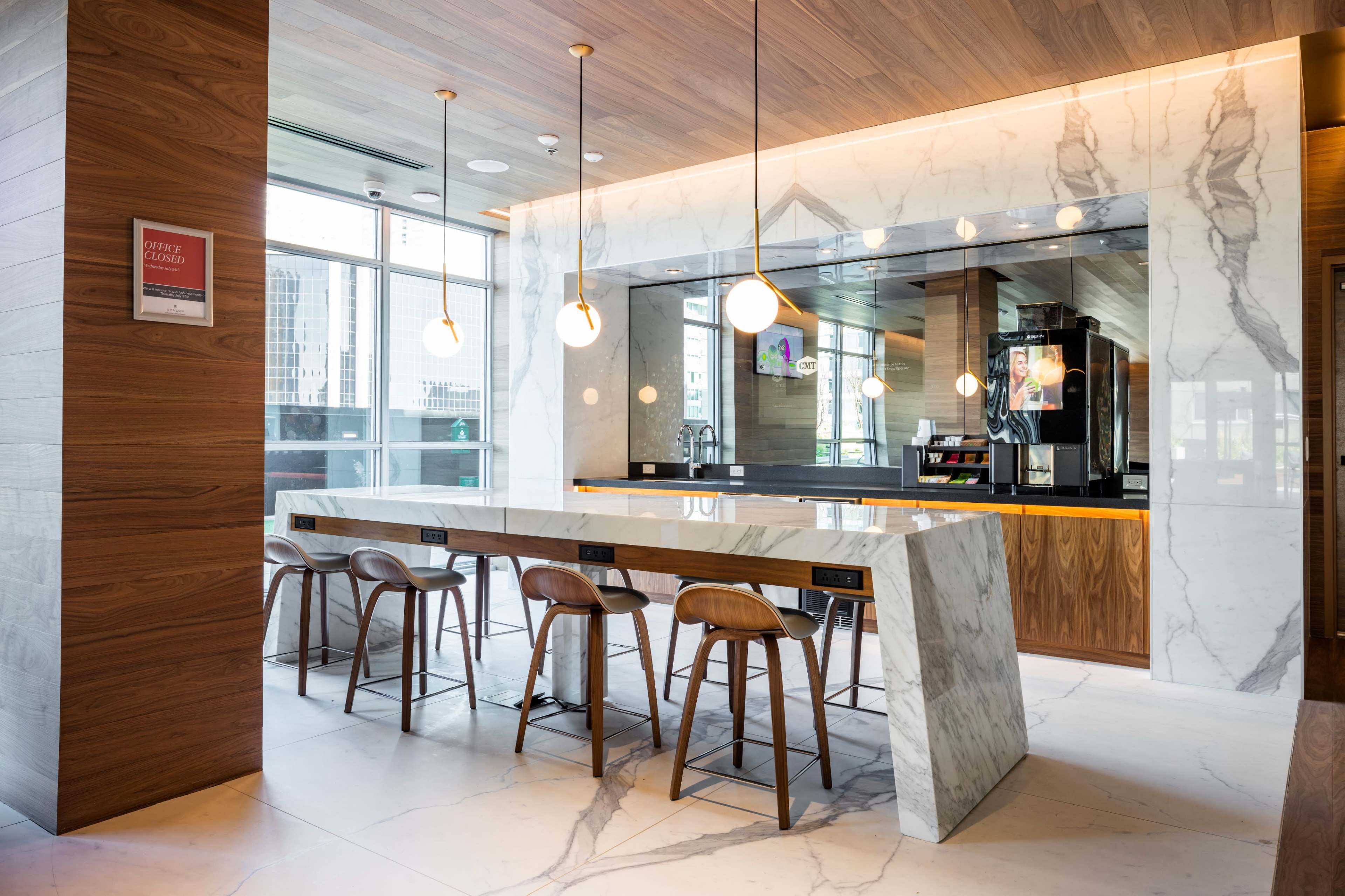 A modern, minimalist kitchen features a marble-topped bar with wooden stools and large windows providing natural light.