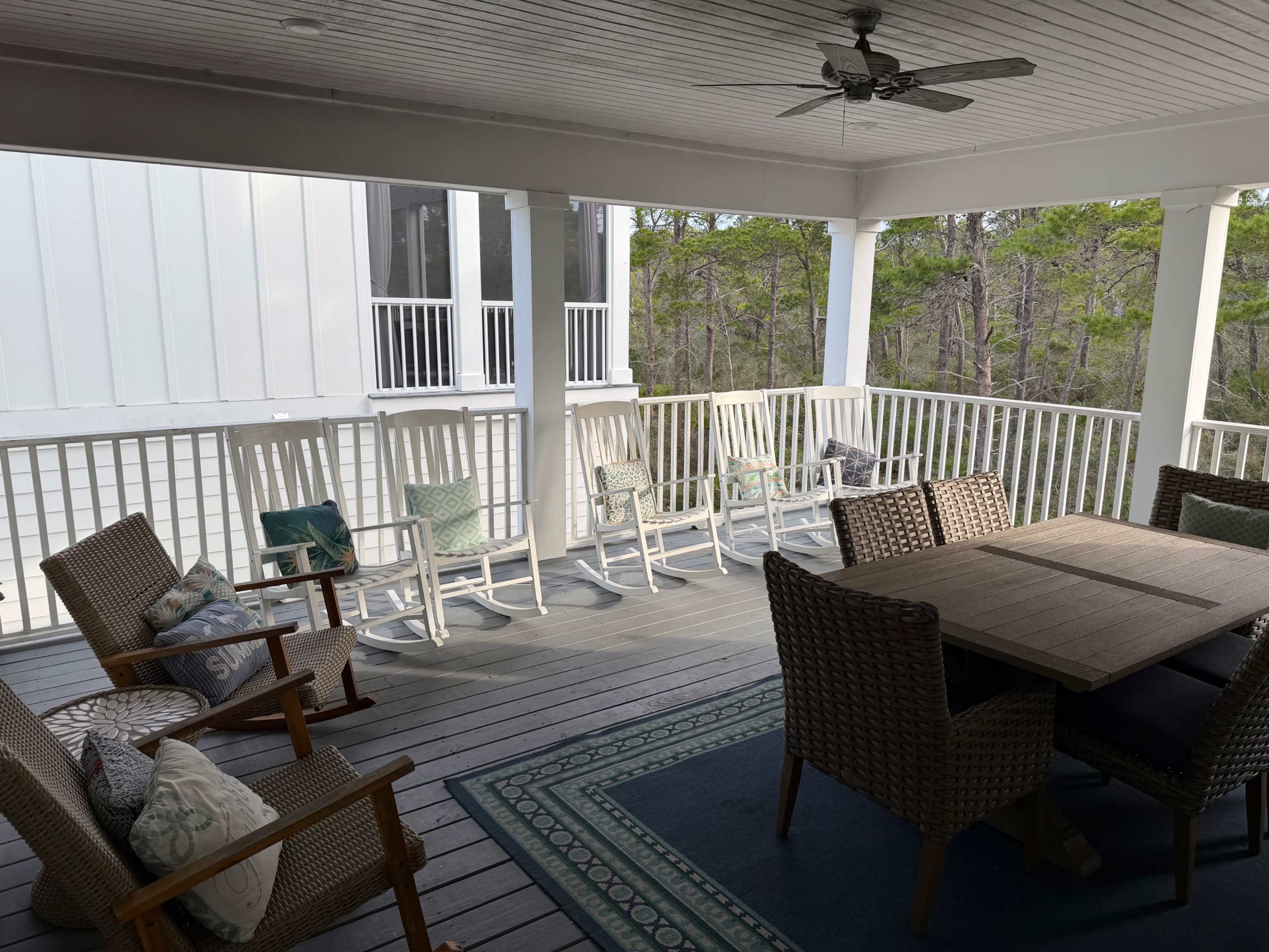 The image shows a spacious porch with several rocking chairs and a dining table surrounded by trees.