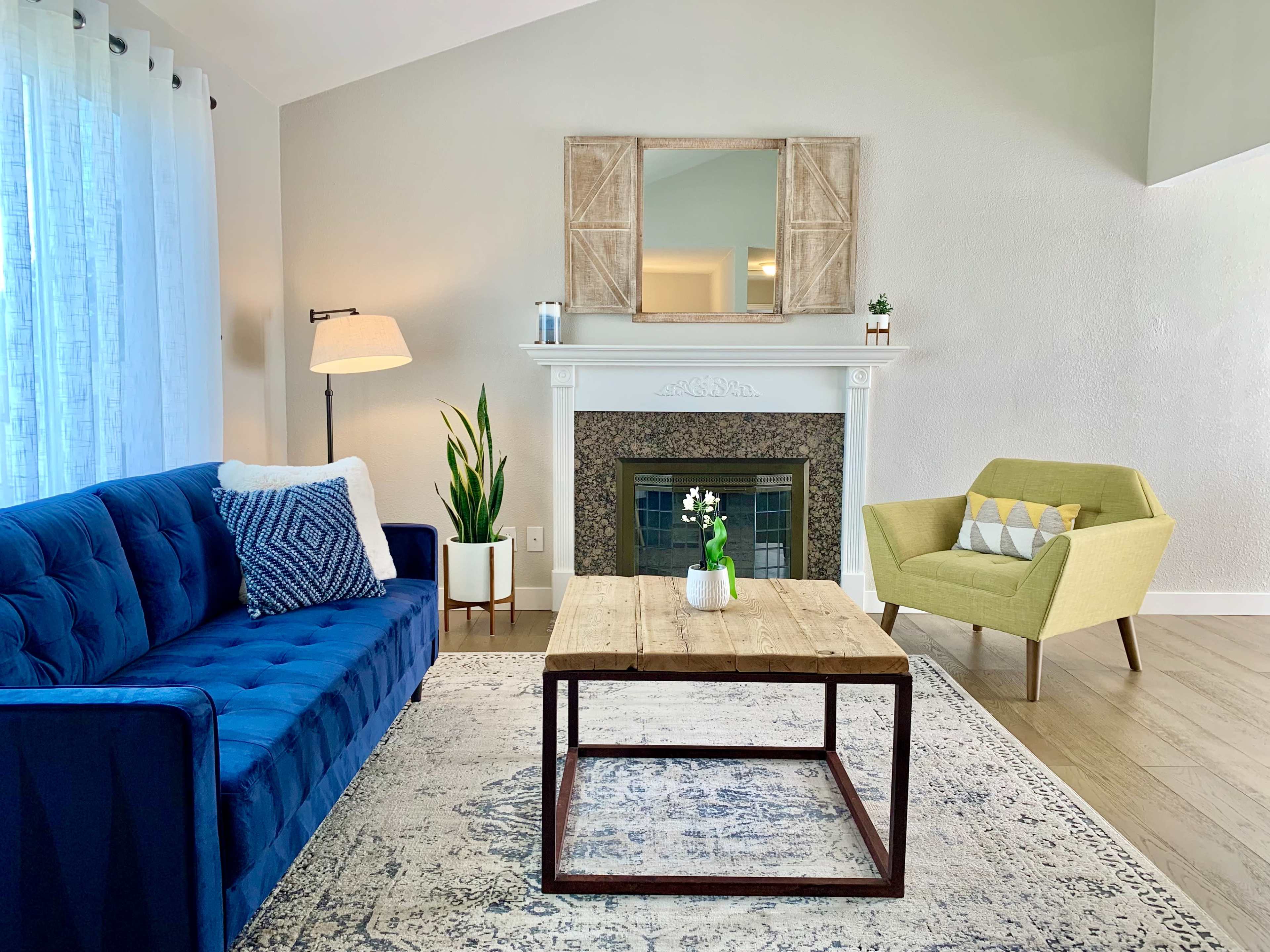 A cozy living room features a blue sofa, a wooden coffee table, a yellow armchair, and a fireplace adorned with a decorative mirror and plants.