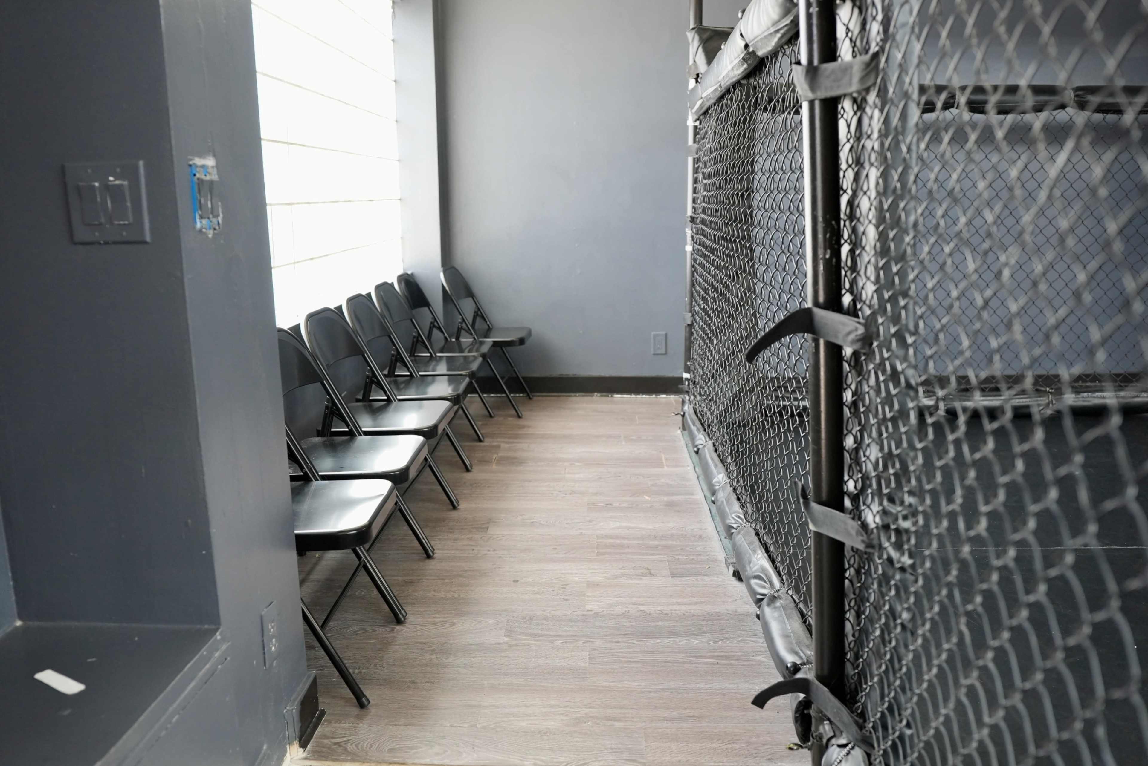 The image shows a narrow space featuring a row of black folding chairs along one wall and a section of a chain-link fence on the opposite side.