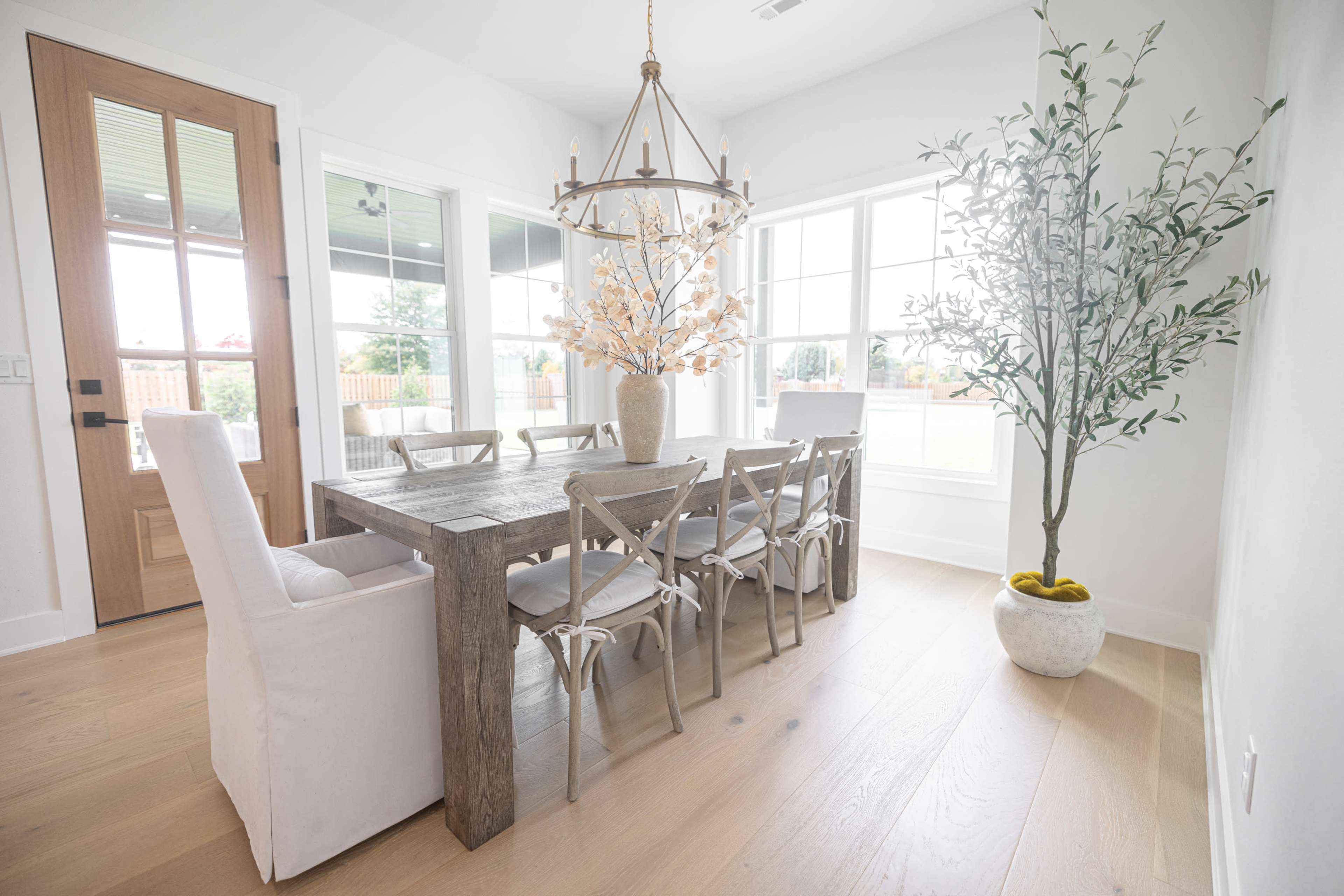A dining area features a wooden table surrounded by chairs, a decorative plant in a pot, and a large window allowing natural light to enter.