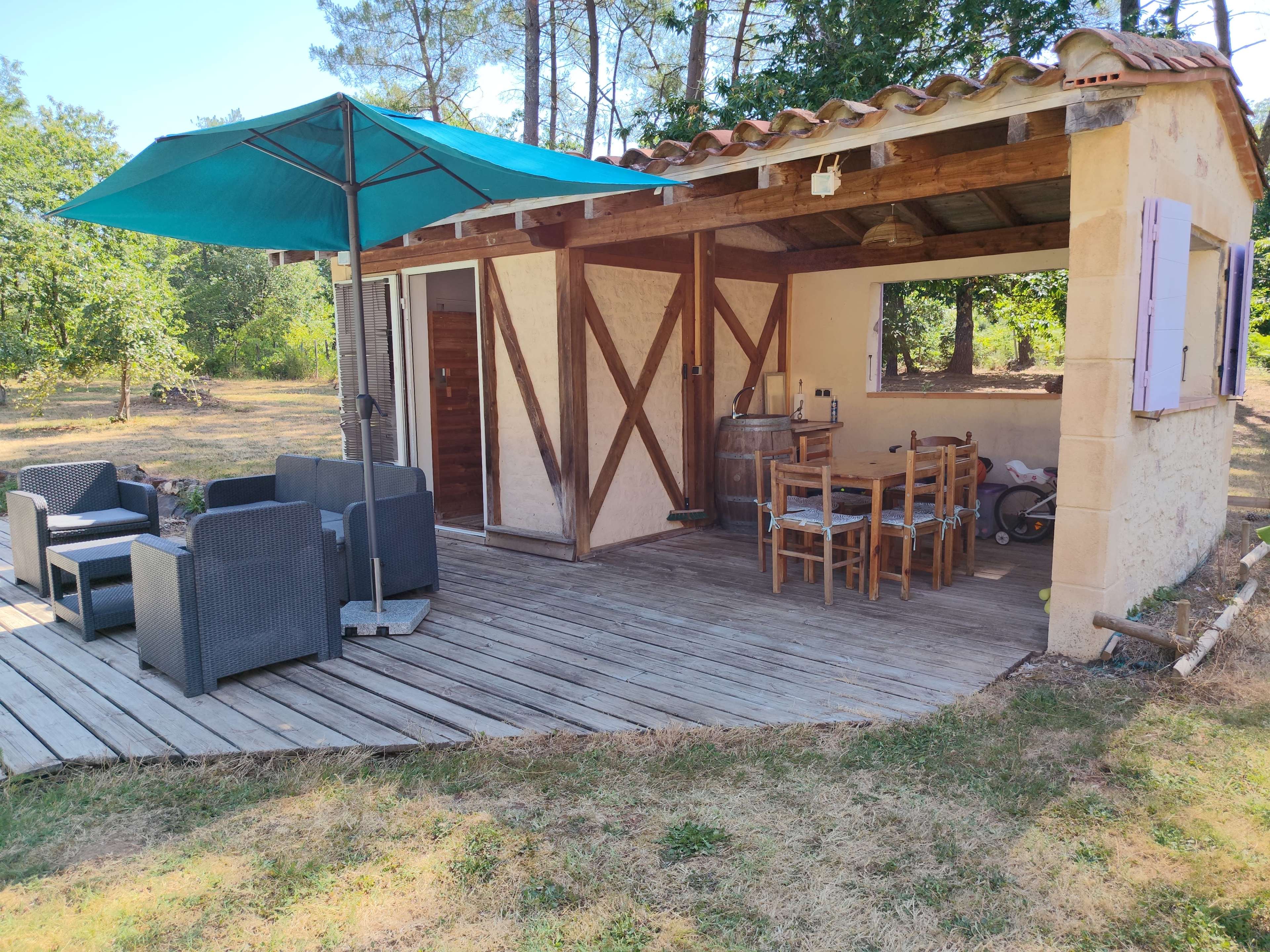 The image shows a small rustic cabin with a wooden deck, equipped with patio furniture and an umbrella, set in a grassy area surrounded by trees.