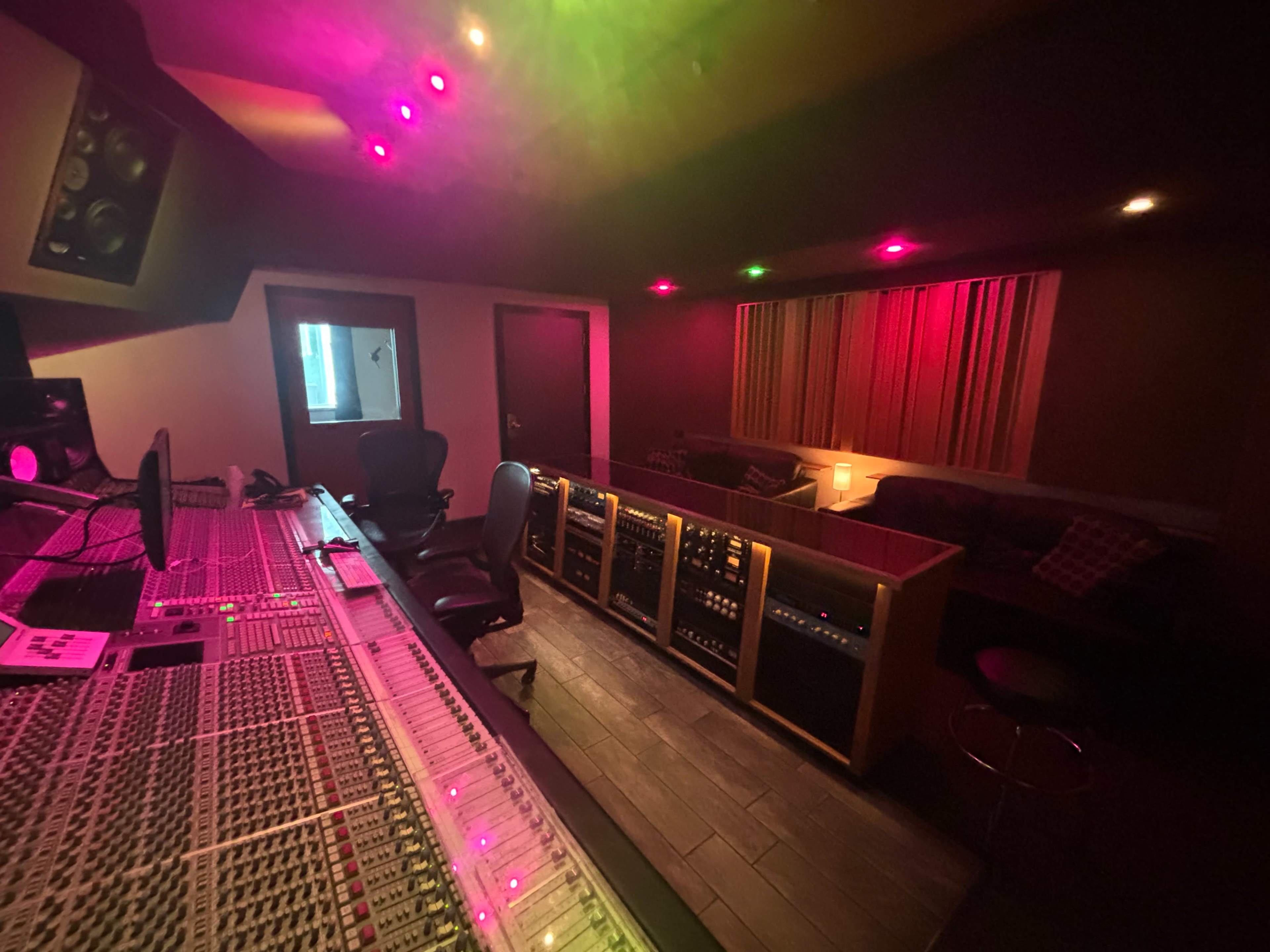 The image shows a dimly lit music recording studio with a large sound mixing console in the foreground and a couch in the background.