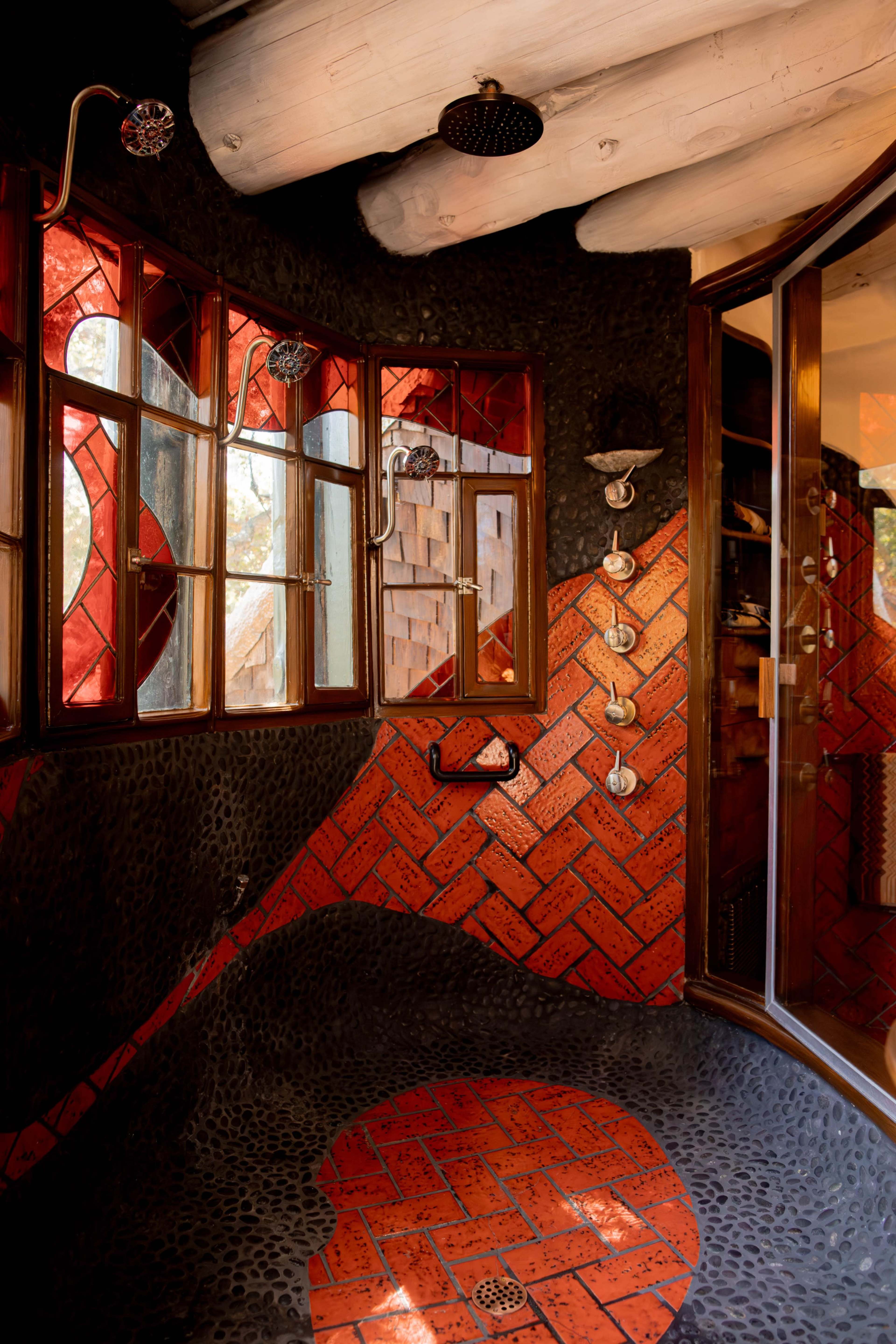 The image shows a uniquely designed bathroom featuring a textured wall with red and black patterns, large windows, and various shower fixtures.