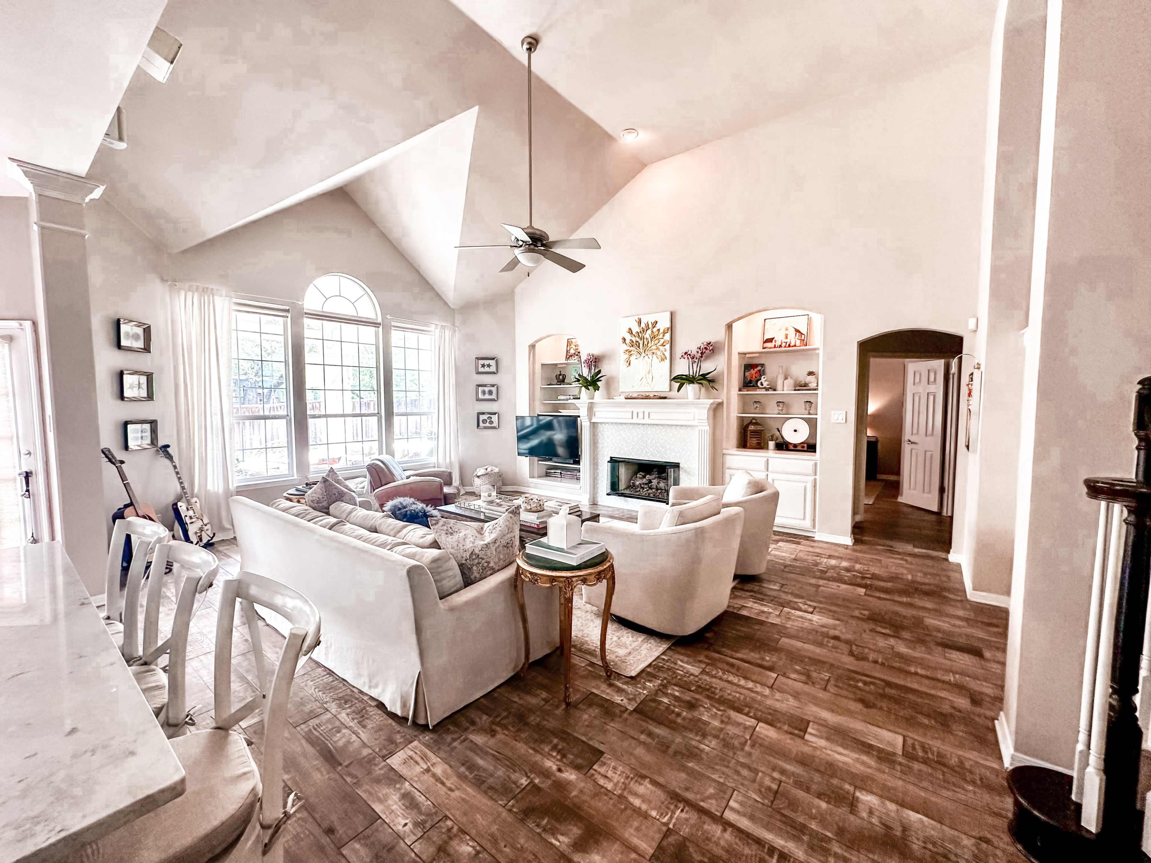 The image shows a spacious living room with high ceilings, a fireplace, and large windows, furnished with a sectional sofa and several decorative elements.