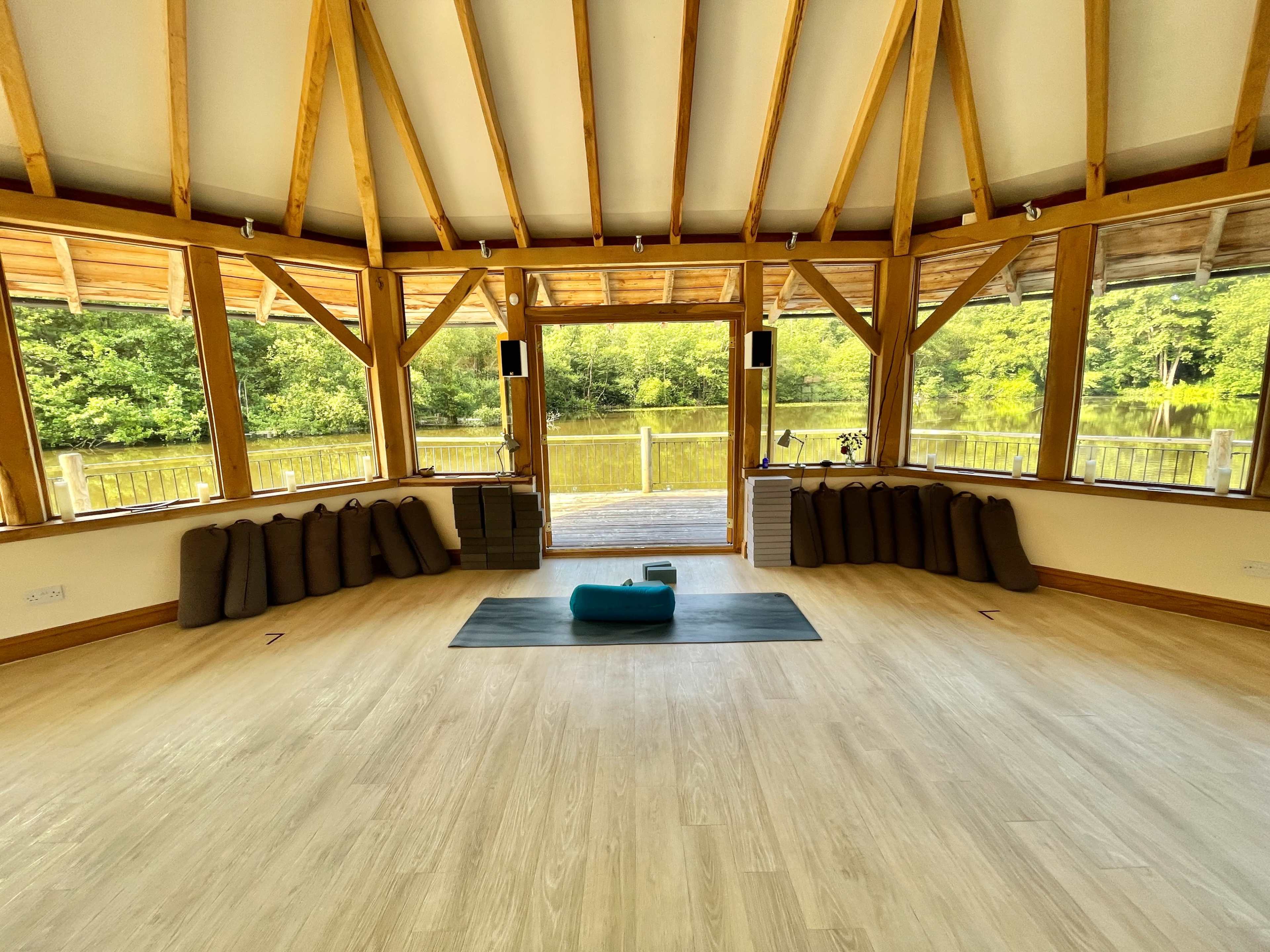 A spacious yoga studio features wooden beams, large windows overlooking a pond, and neatly arranged yoga mats and props.