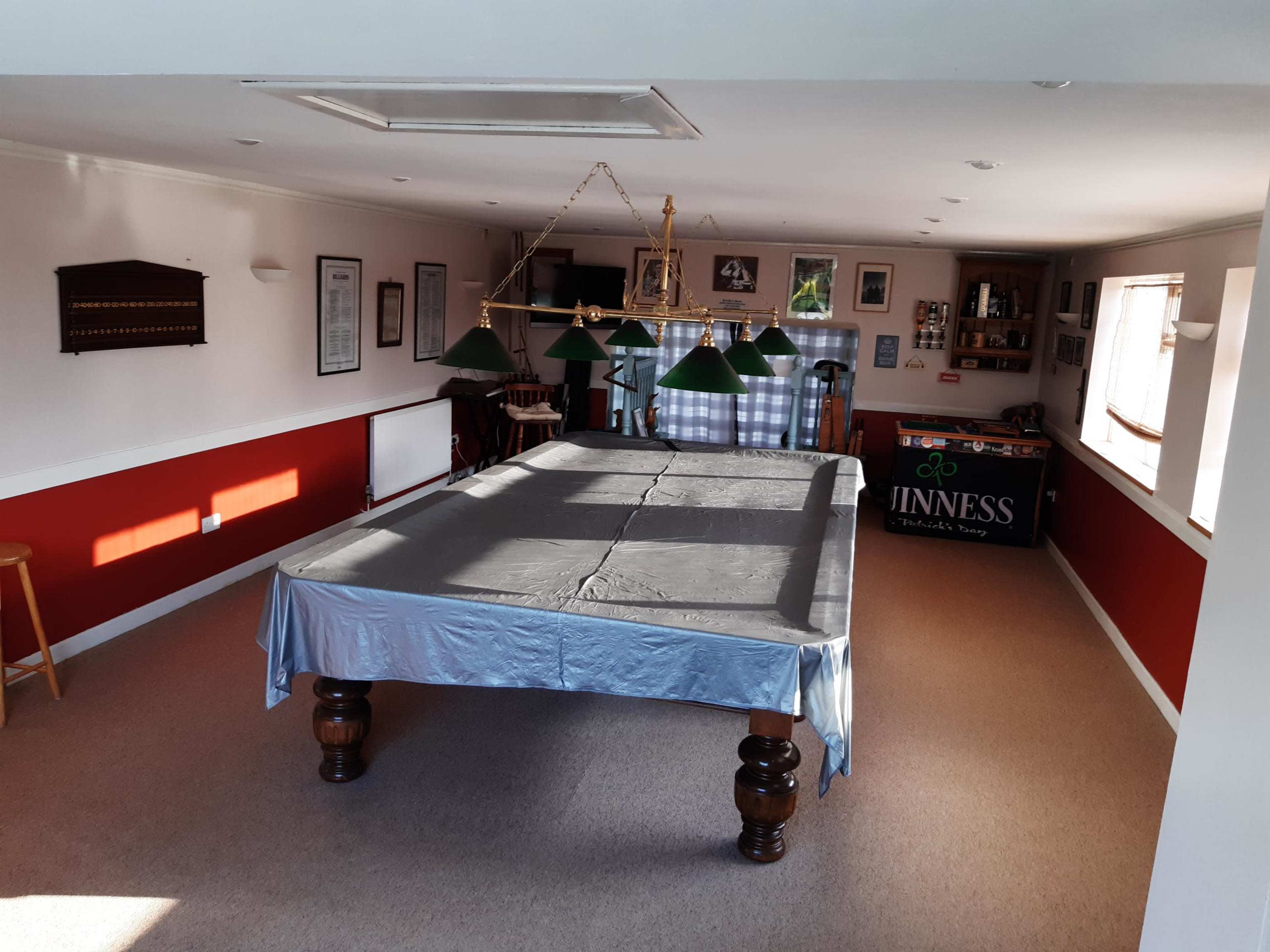 The image shows a spacious game room featuring a billiards table with a gray cover, green pendant lights overhead, and a bar area displaying a Guinness advertisement.