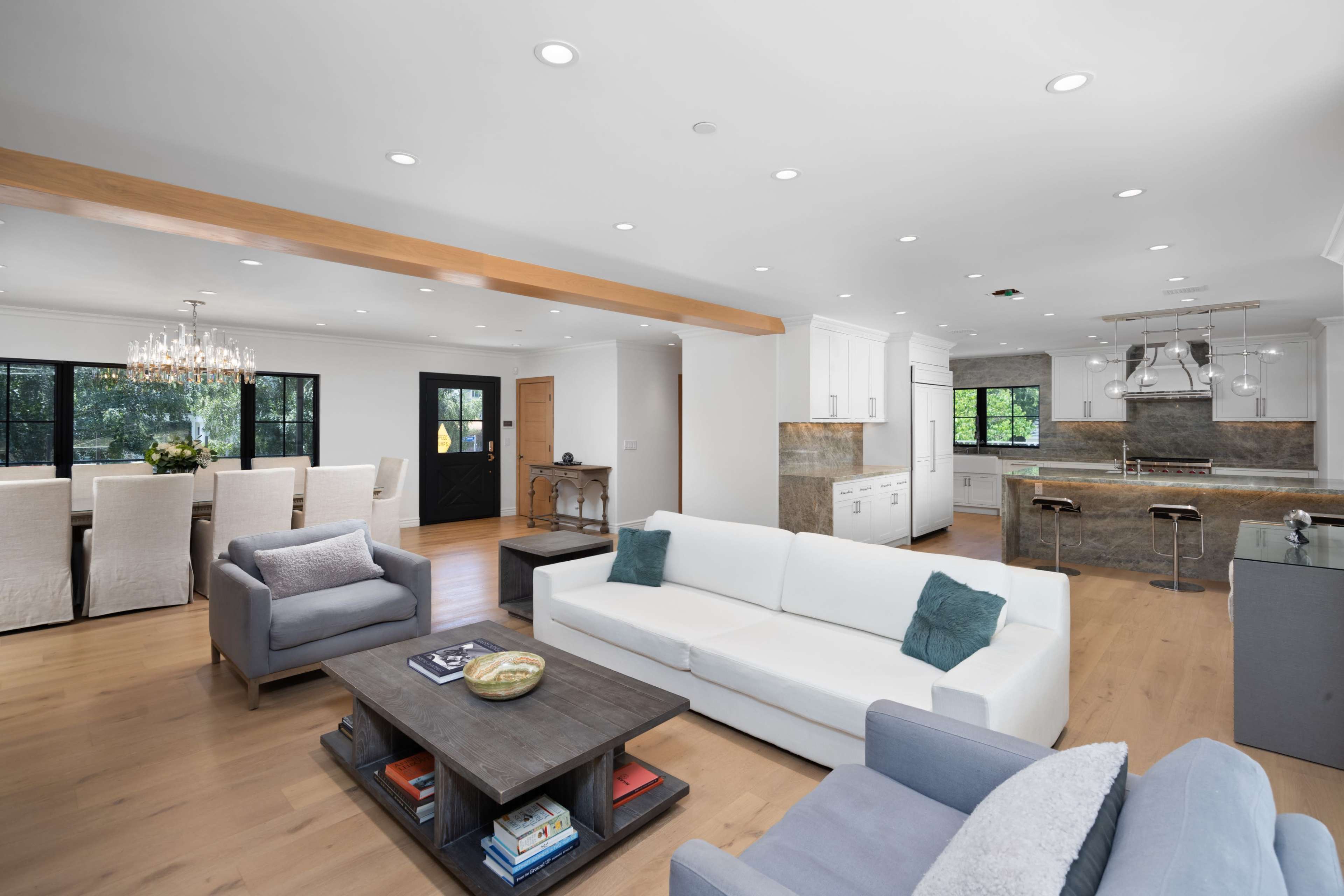 A modern, open-concept living area features a white sofa, a light gray armchair, a coffee table with decorative items, and a kitchen with bar stools in the background.