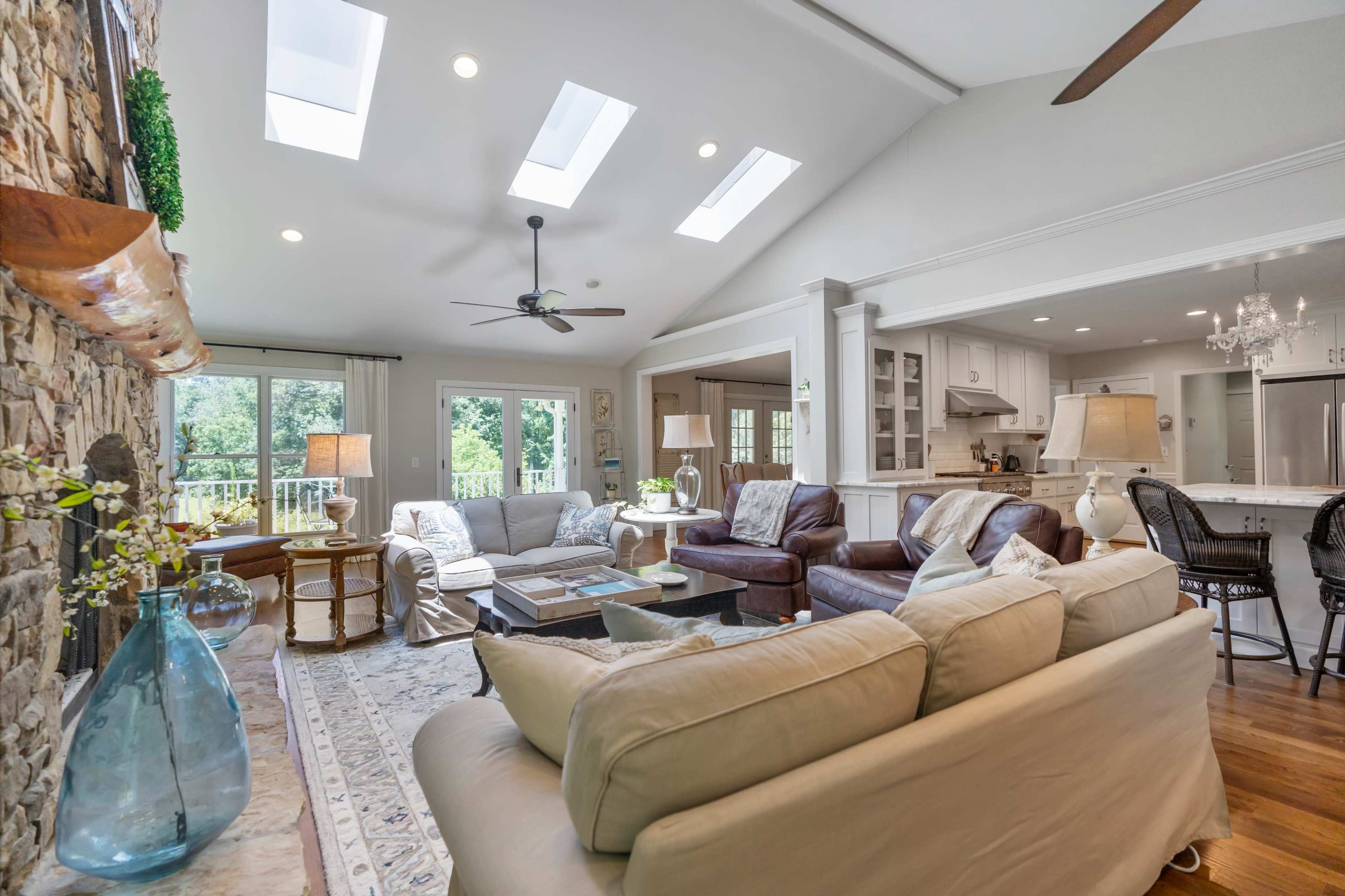 A spacious living room with large windows, a stone fireplace, and a combination of light-colored and dark brown furniture, leading into a bright kitchen area.