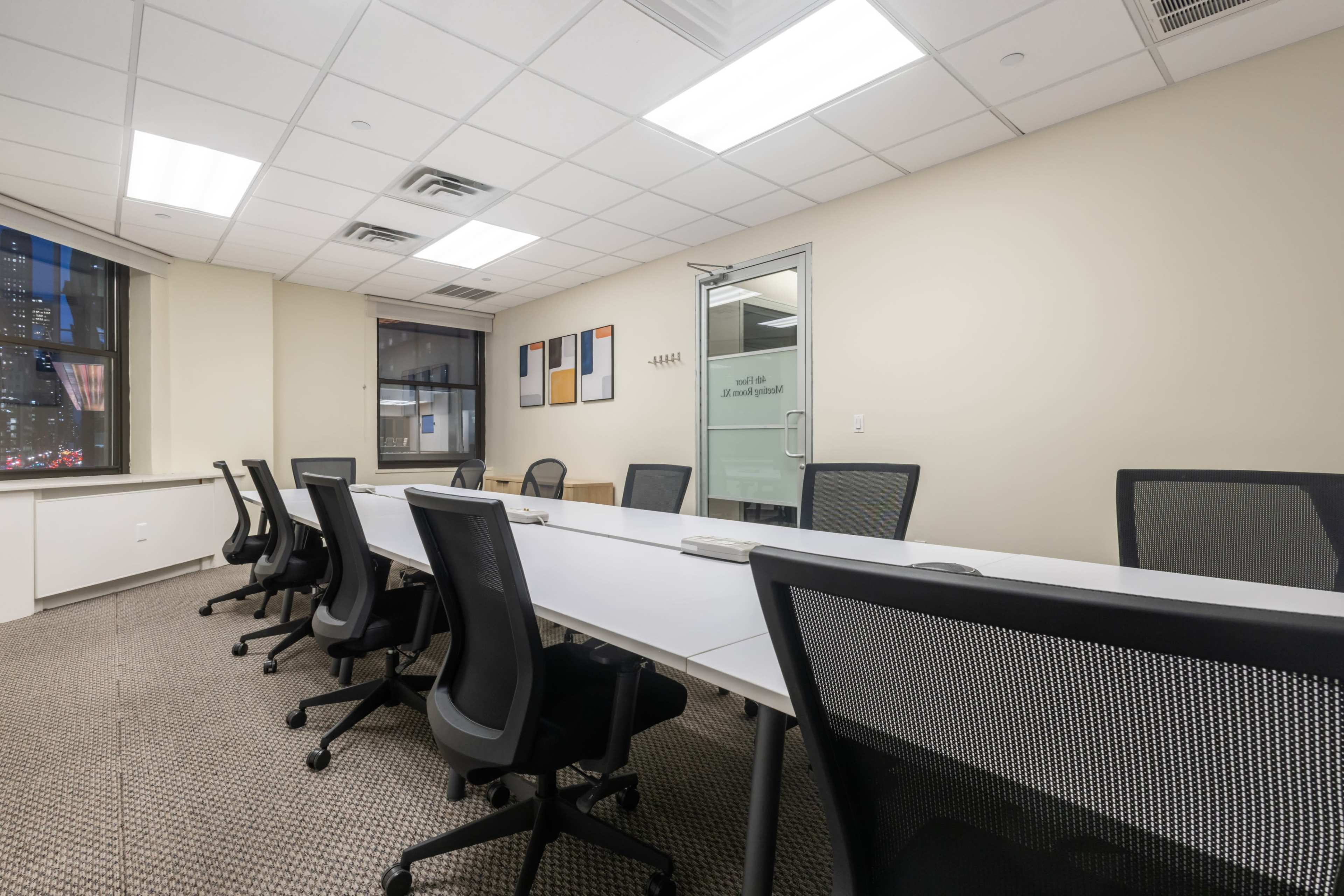 A modern conference room features a long white table surrounded by black office chairs, with large windows and minimalist decor.