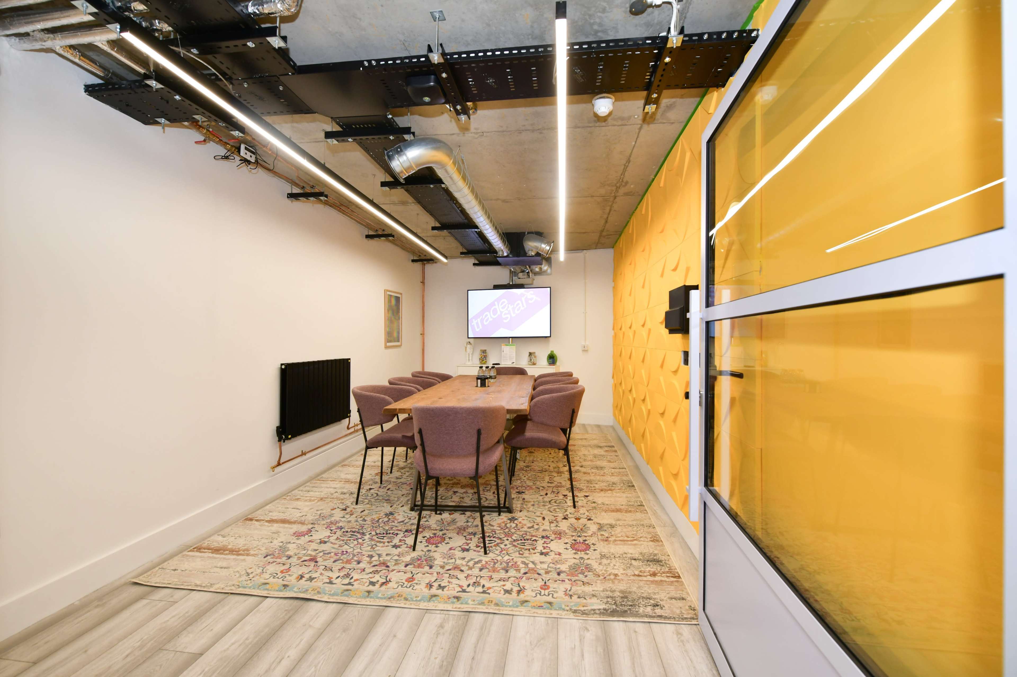 A conference room features a long wooden table surrounded by purple chairs, with a large screen and yellow wall panels.