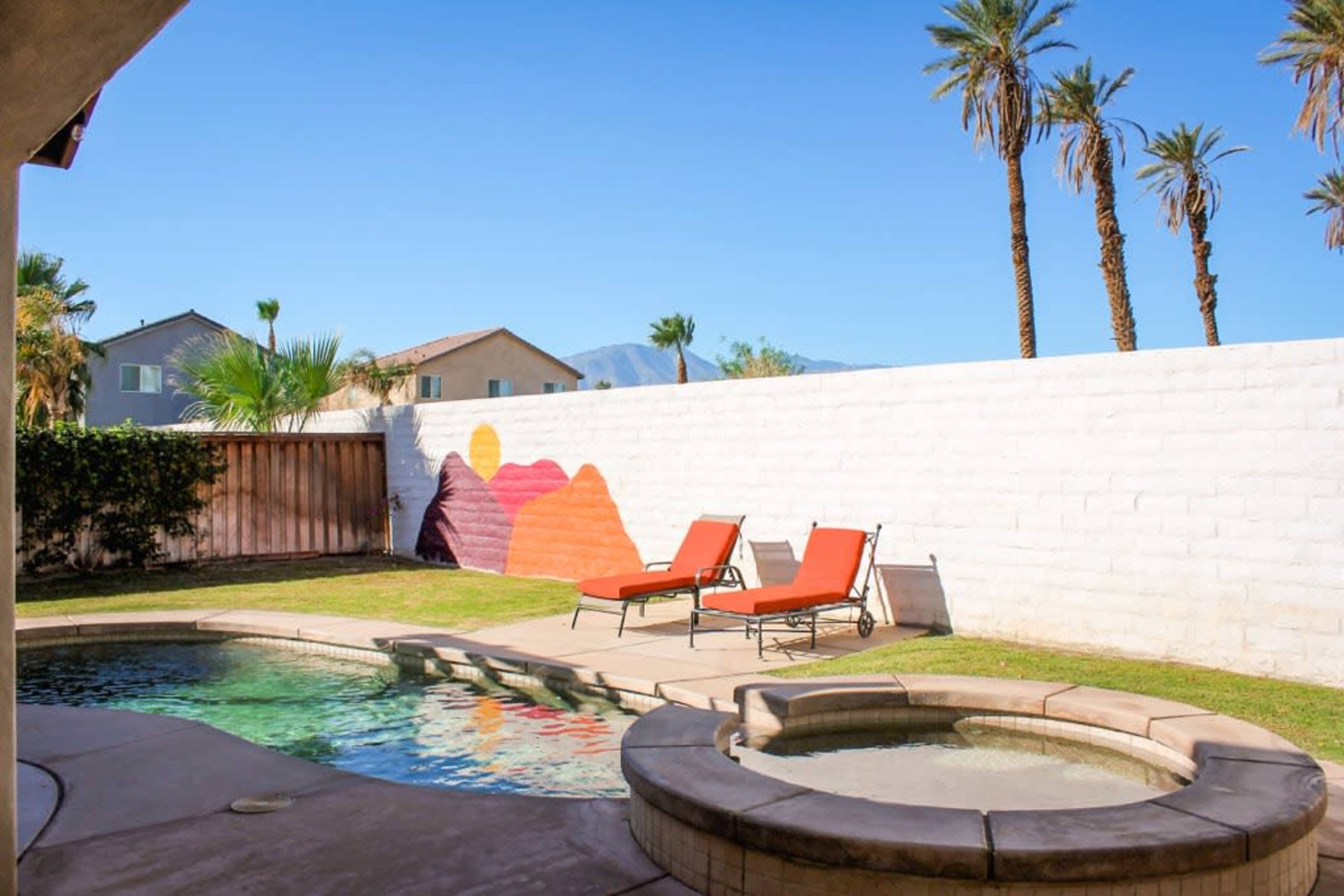 A backyard with a swimming pool, a spa, and two orange lounge chairs beside a wall featuring a colorful mural, with palm trees in the background.