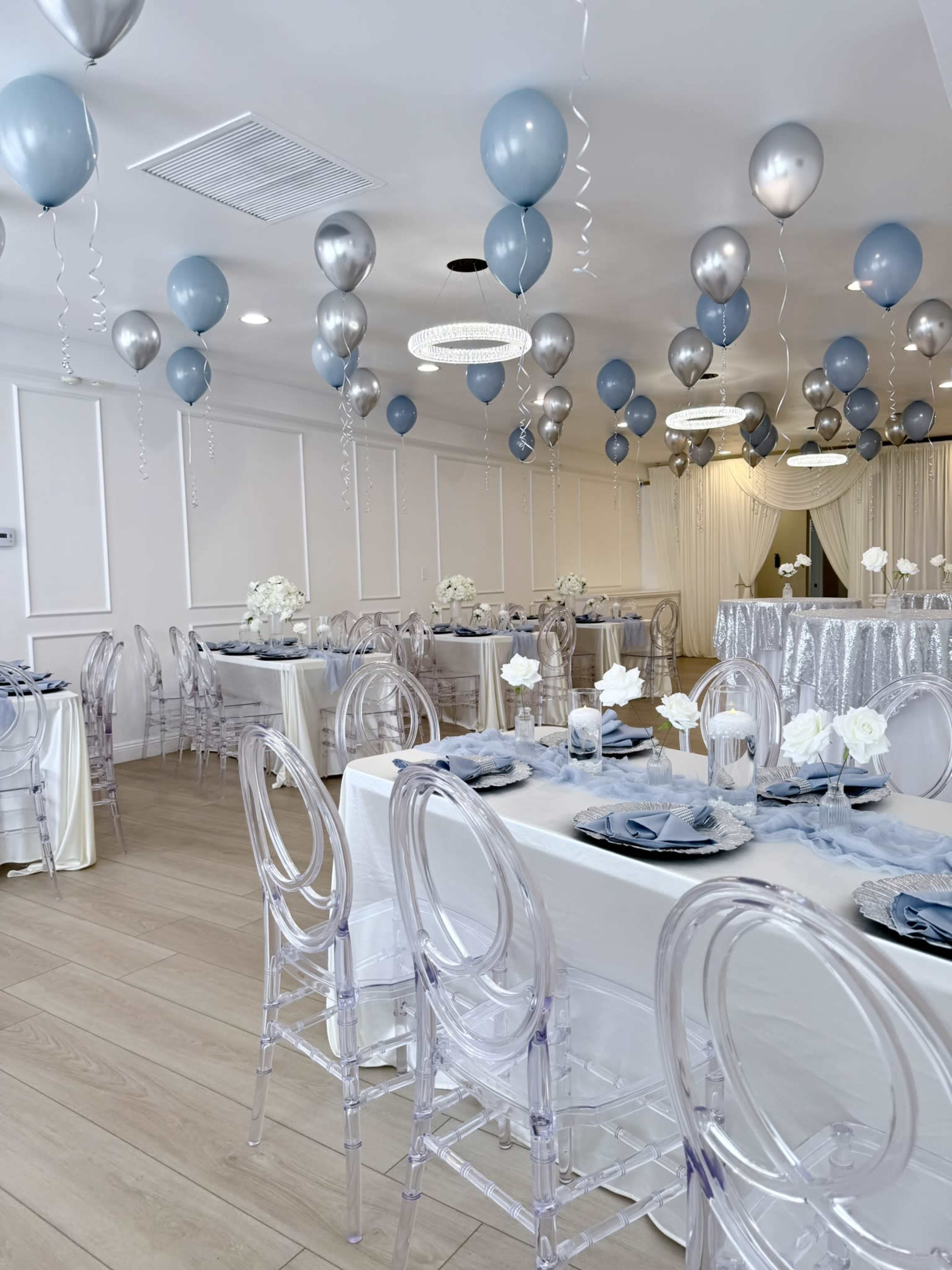 A decorated event space features tables set with blue plates and white flowers, surrounded by silver and blue balloons hanging from the ceiling.
