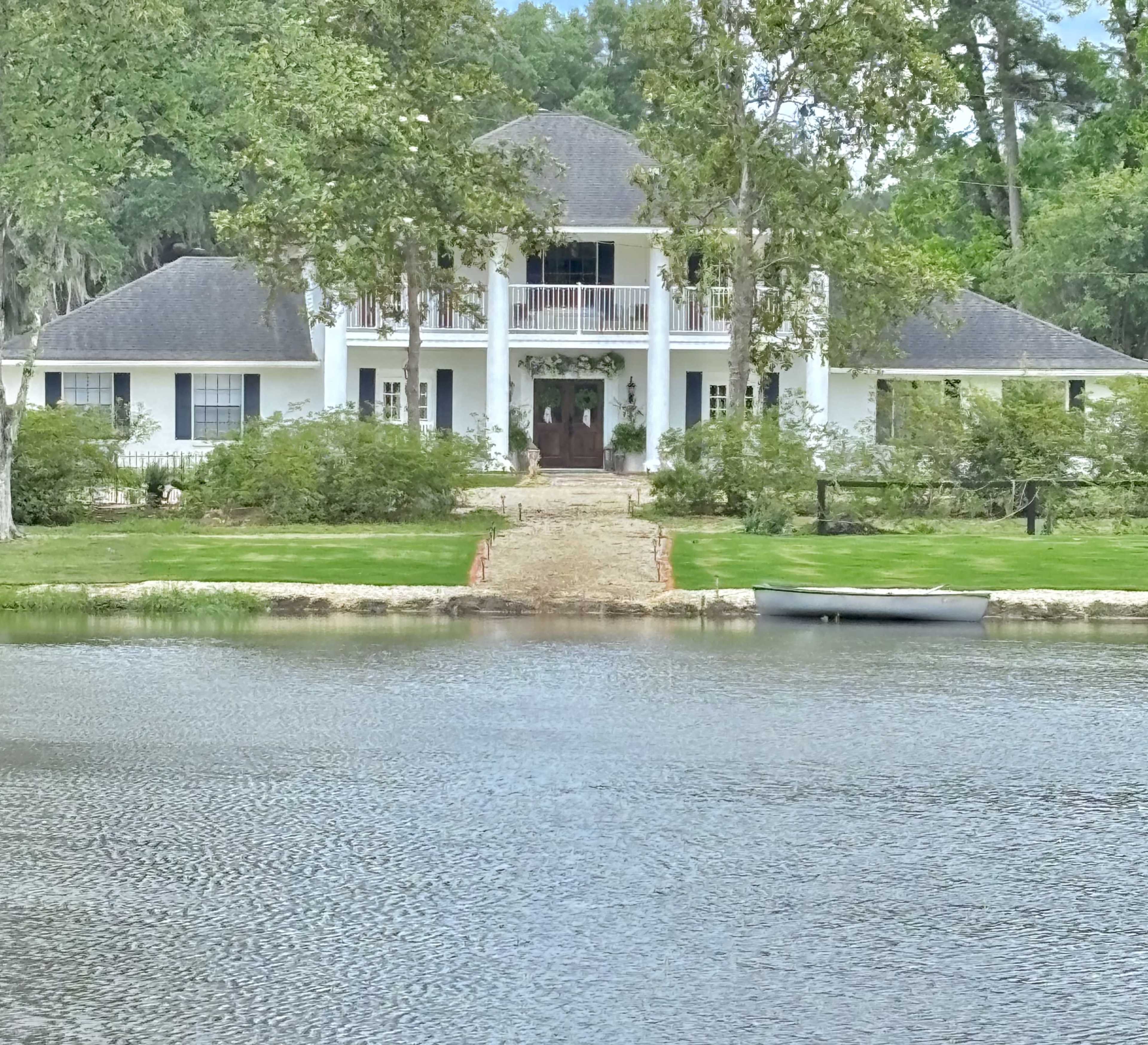 The image shows a large white house with a landscaped front yard and a body of water in the foreground.