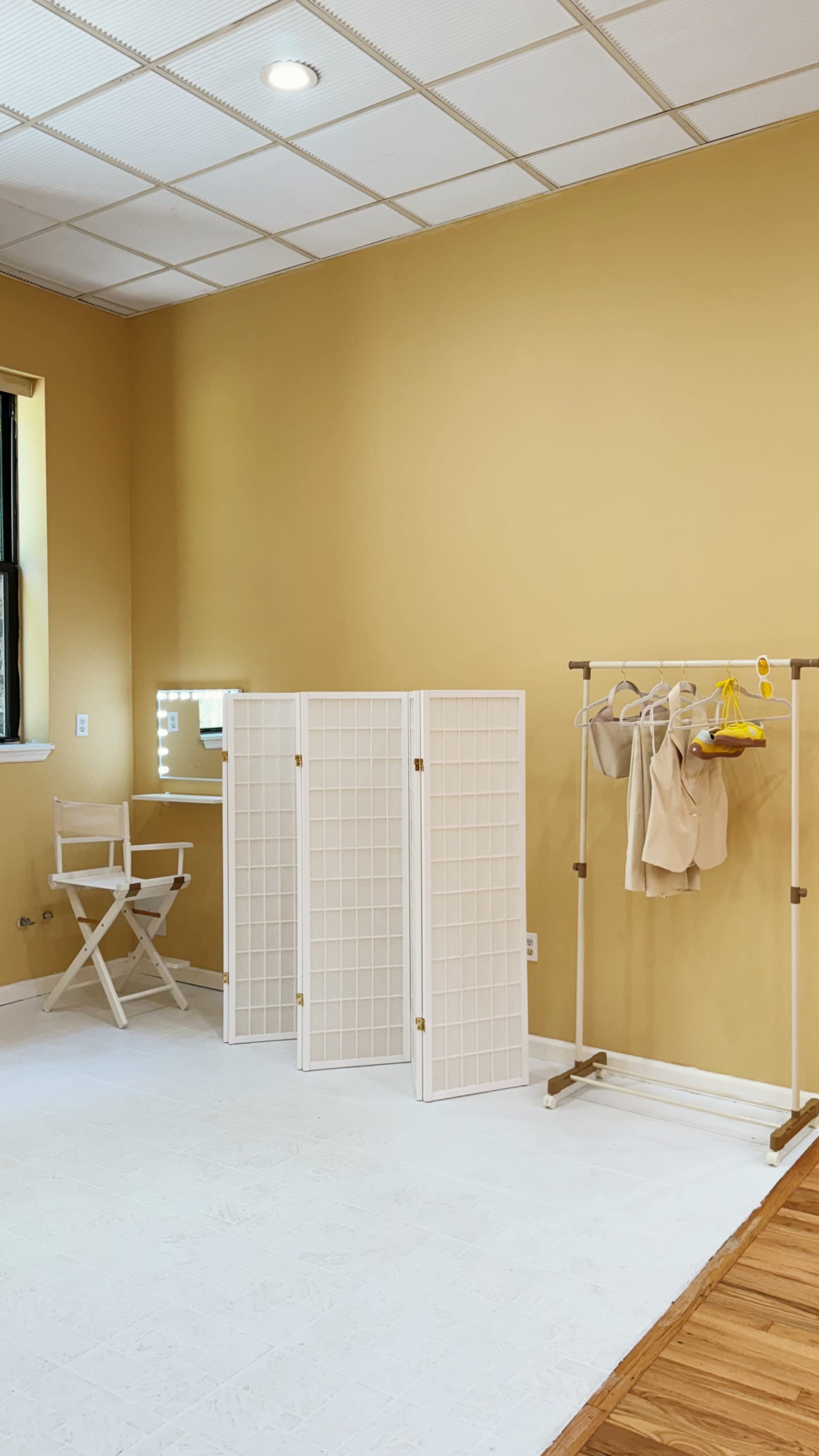The image shows a room with beige walls, a folding screen, clothing rack with garments, a director's chair, and a lighted mirror.