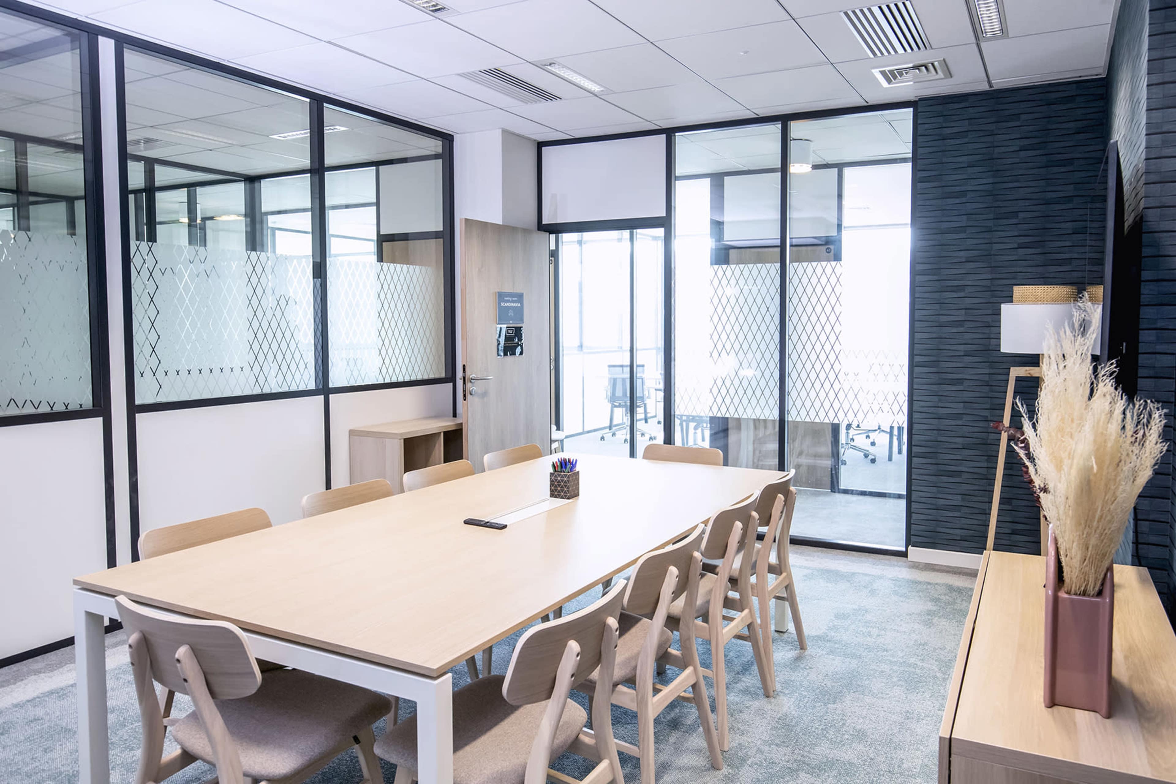 A modern conference room features a large wooden table surrounded by chairs, with glass walls providing views into adjacent rooms.