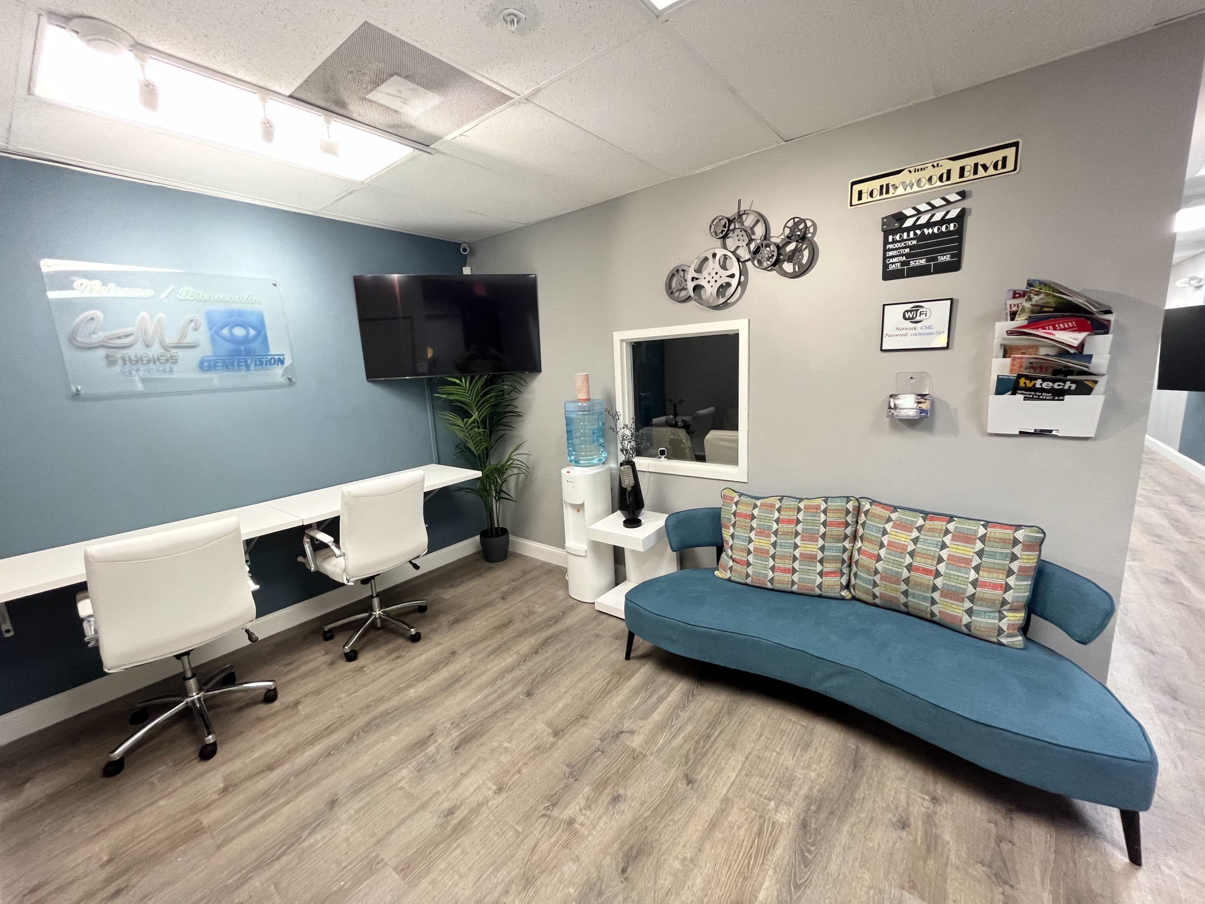 The image shows a modern office space with a teal accent wall, two white desks with chairs, a water cooler, and a colorful couch against the wall.