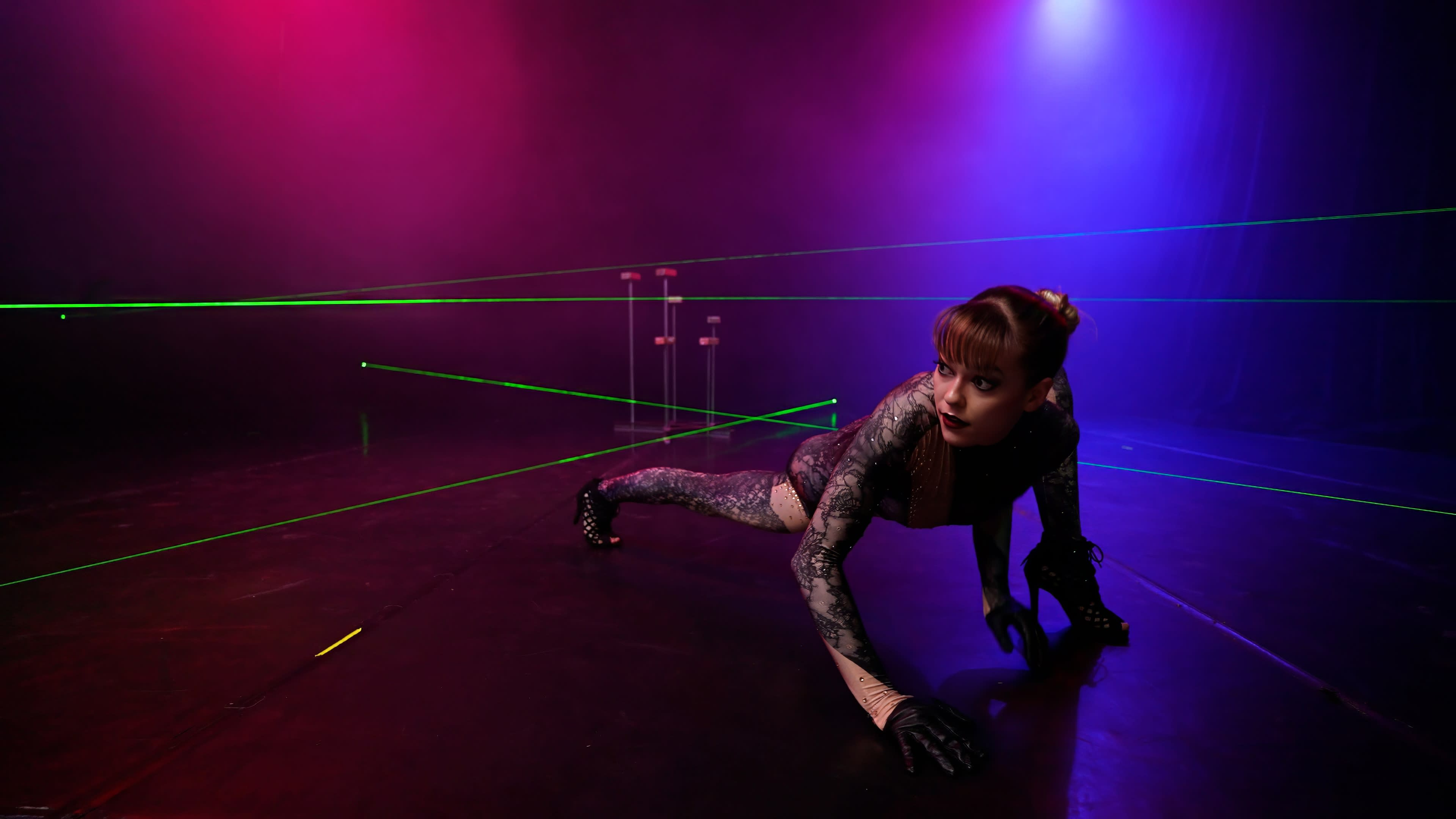 A performer in a cat-like pose navigates a stage filled with green laser beams against a backdrop of purple and blue lighting.