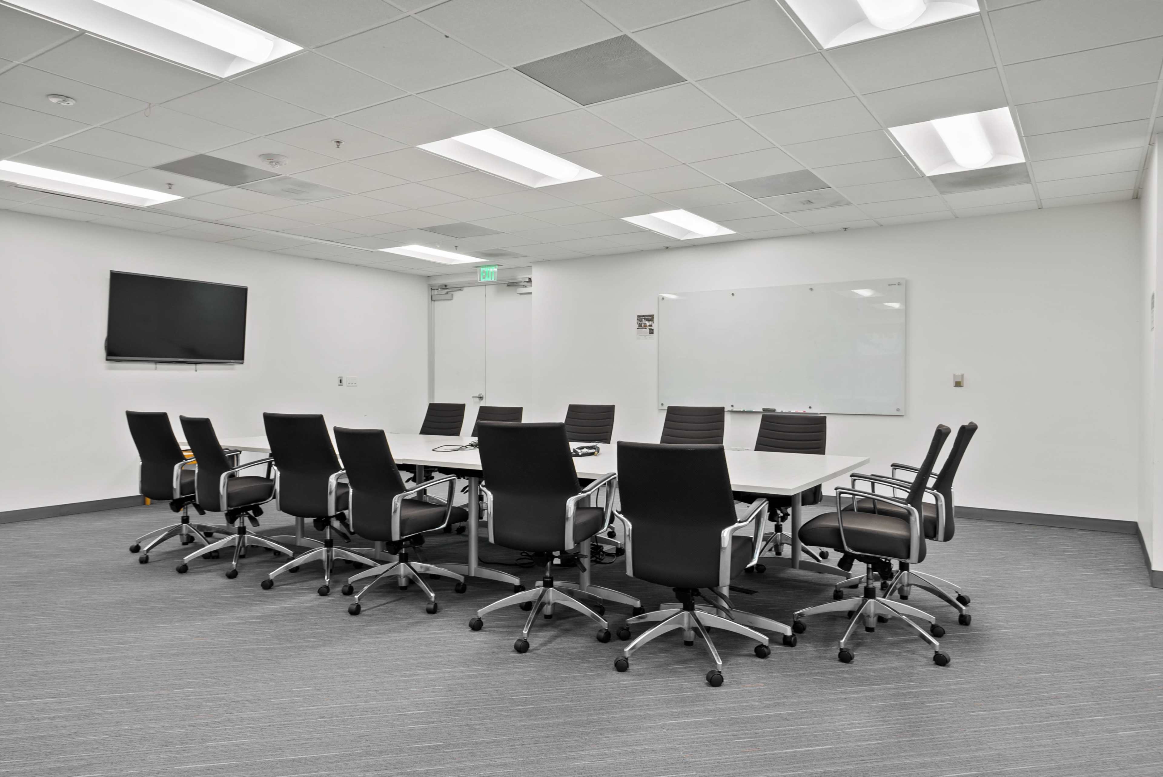 A modern conference room features a long table surrounded by ten ergonomic chairs, with a whiteboard and television mounted on the walls.