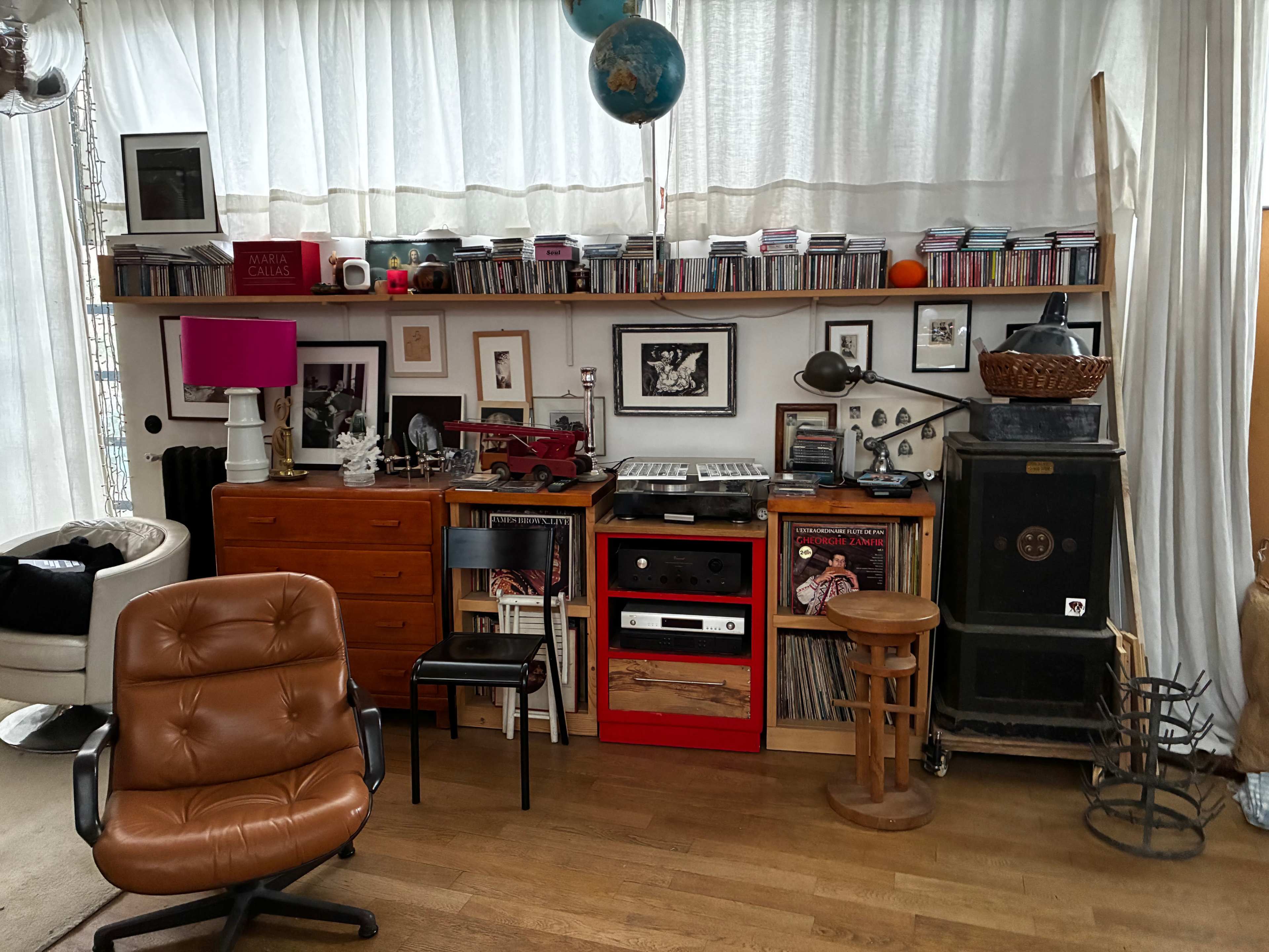 The image shows a living space with a collection of CDs on a shelf, framed pictures on the wall, and various pieces of furniture, including a chair, a stool, and a red media unit.