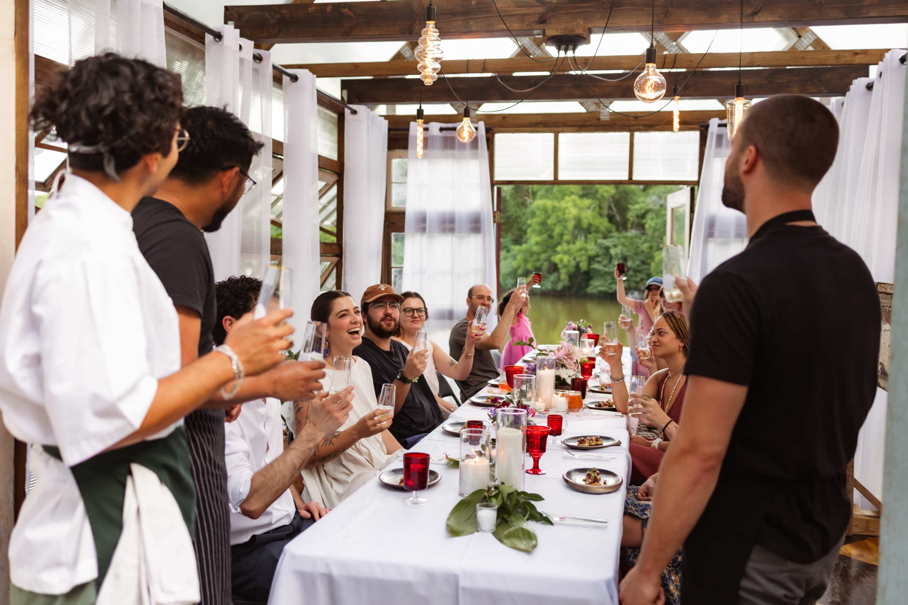 A group of people cheers with drinks at a long table set in a bright, open space with large windows.