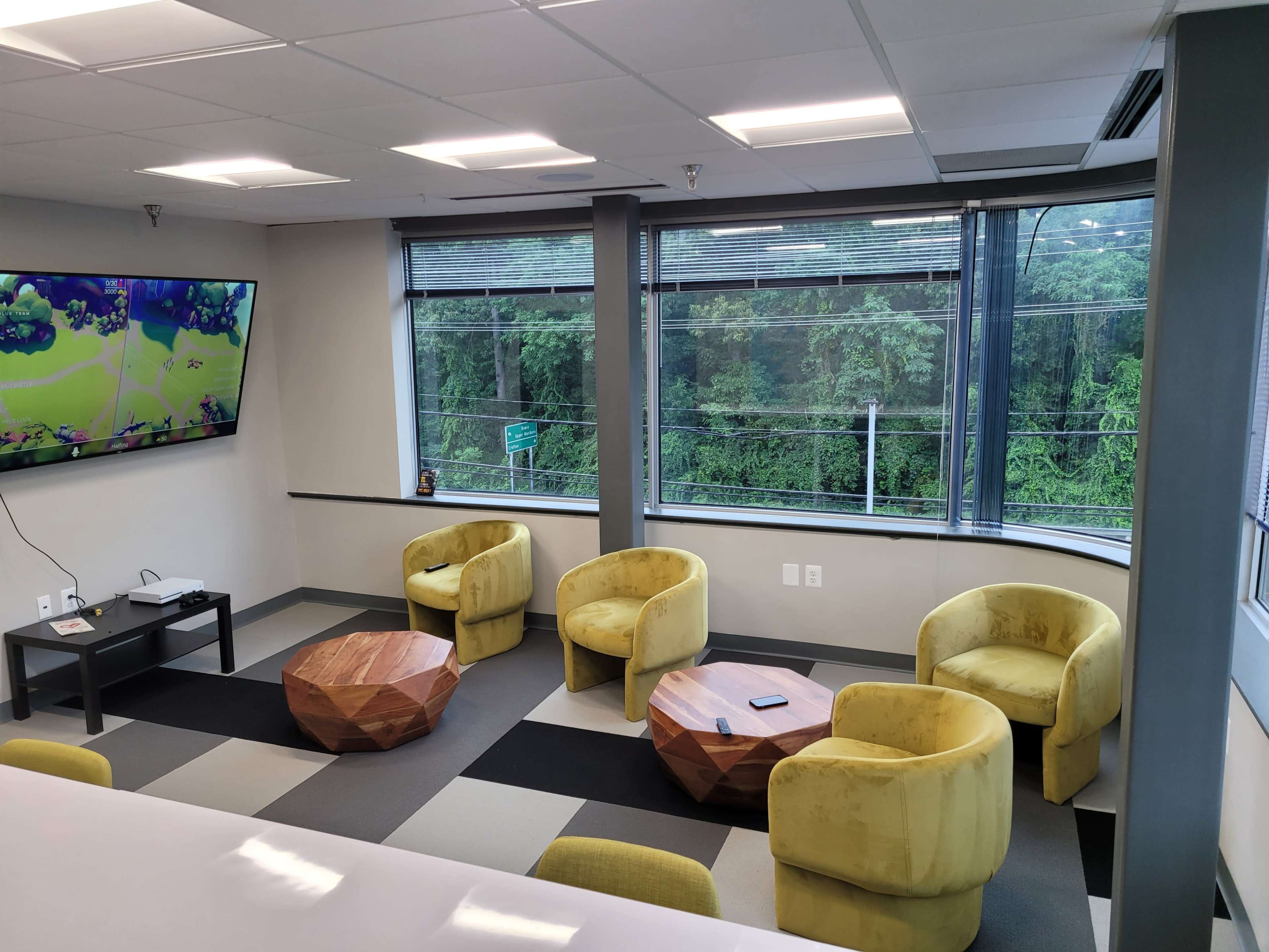 A modern lounge area with four yellow chairs and two wooden coffee tables, set against a backdrop of large windows overlooking greenery.
