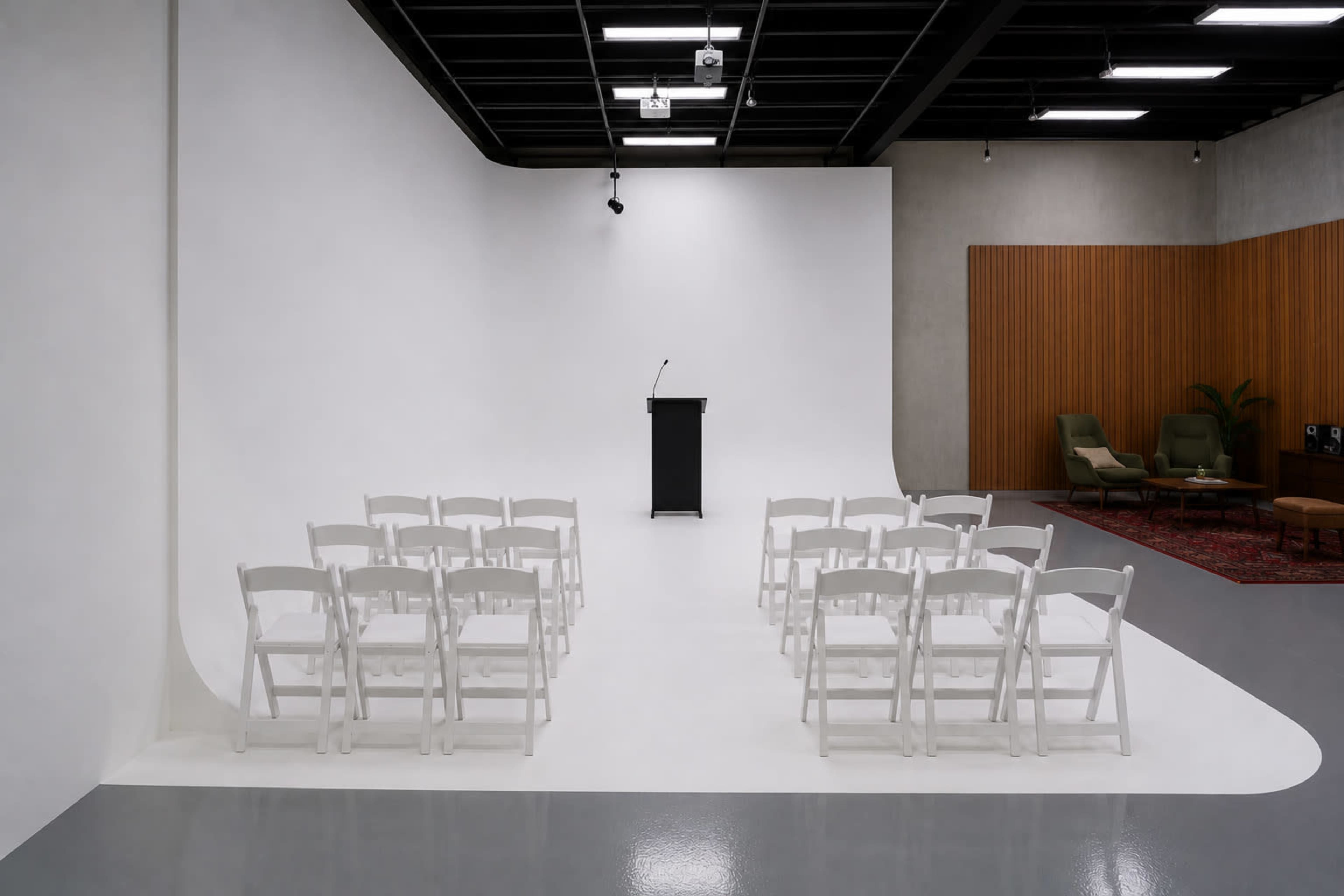 The image shows a minimalist presentation space featuring a black podium and white folding chairs arranged in a semicircle against a plain backdrop.
