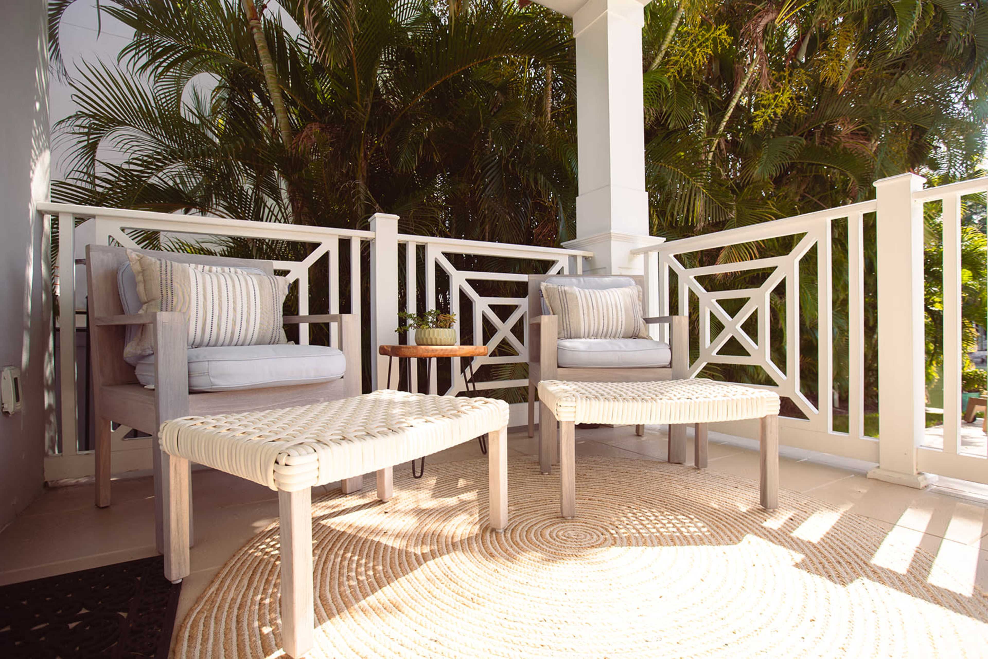 A small balcony features two armchairs with cushions and a round side table, surrounded by lush green plants.