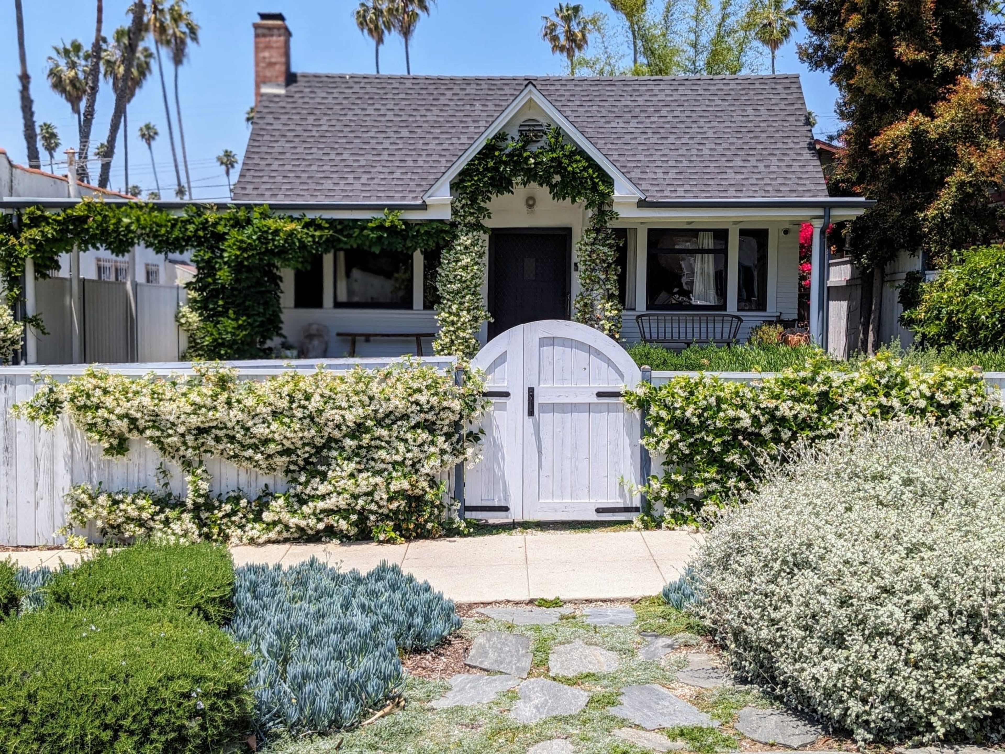 A white picket fence adorned with climbing greenery surrounds a charming, cottage-style house with a gray roof and a well-maintained front yard.