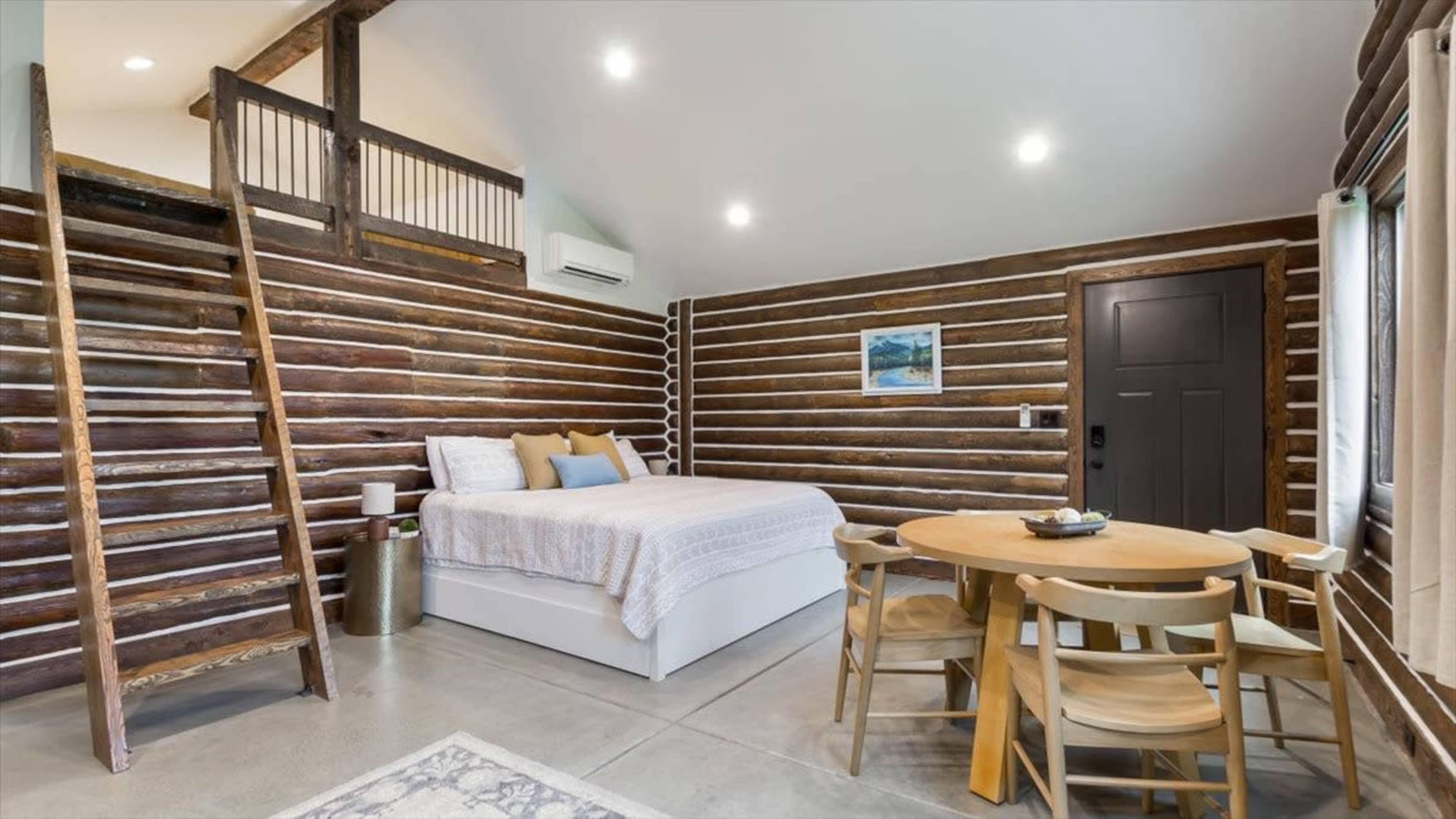 The image shows a compact, modern studio with log cabin walls, a bed, a dining table, and a staircase leading to a loft area.