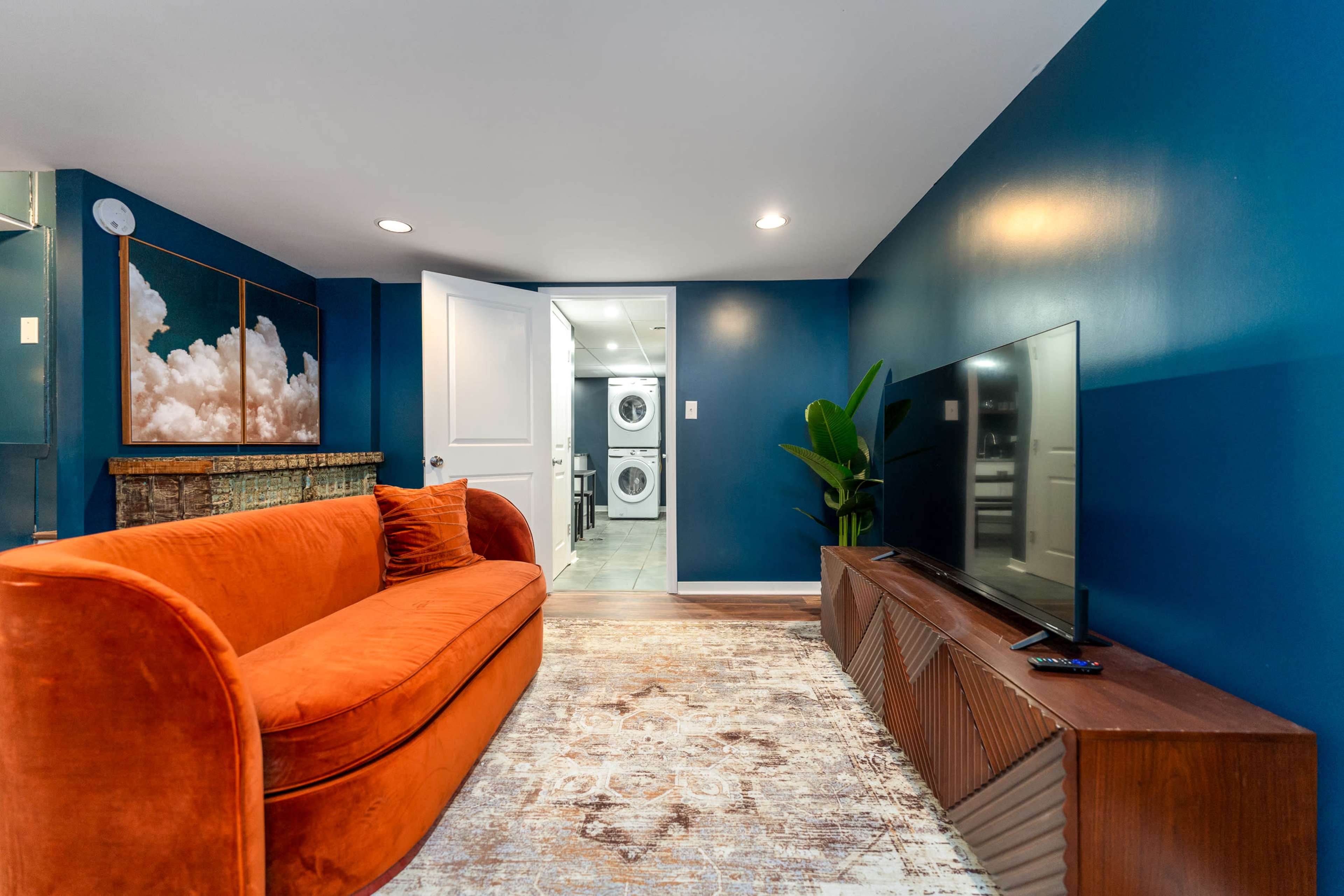 A bright blue room features an orange sofa, a wooden TV stand, and a doorway leading to a laundry area.