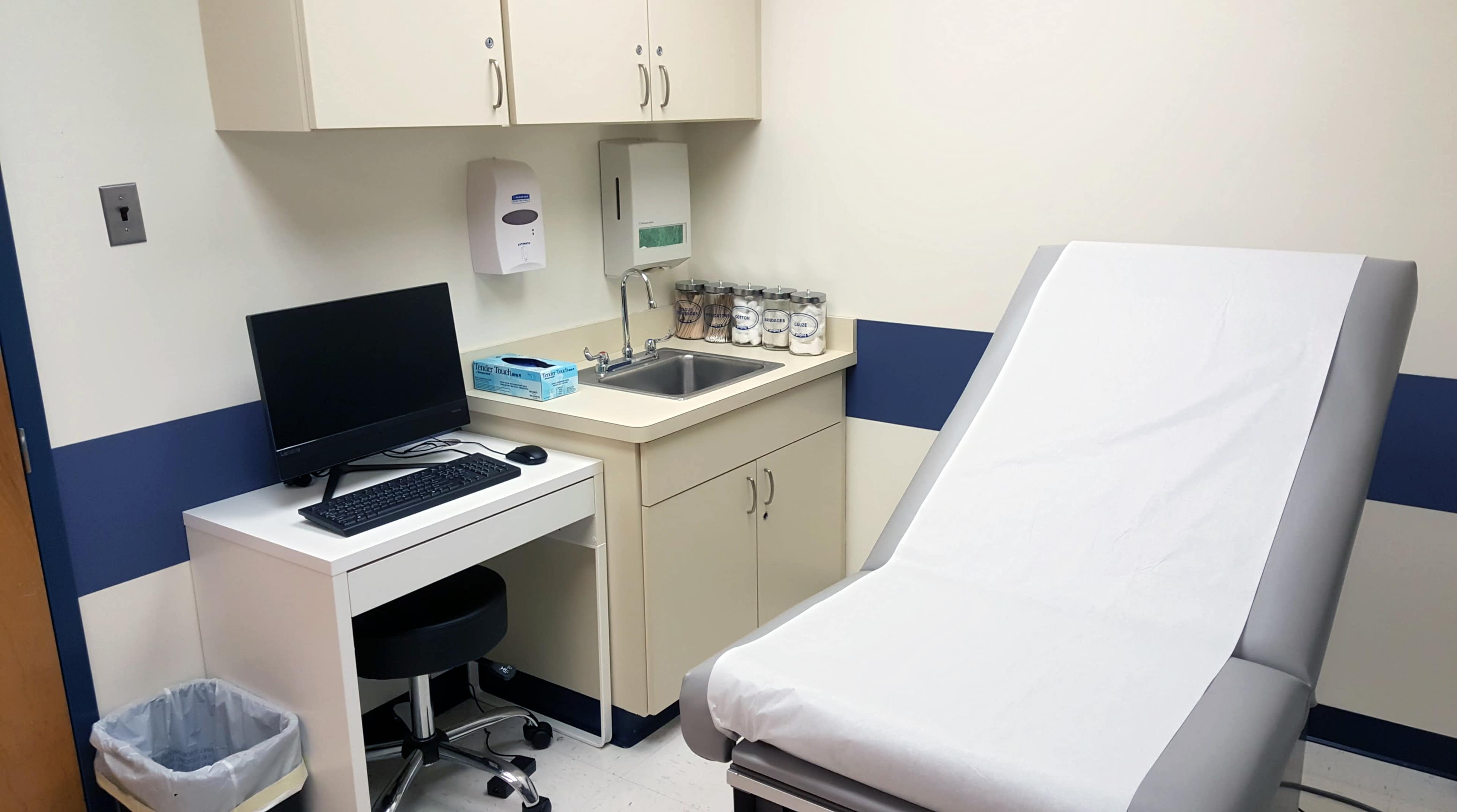 A medical examination room featuring a patient examination table, a desk with a computer, and a sink surrounded by cabinets.
