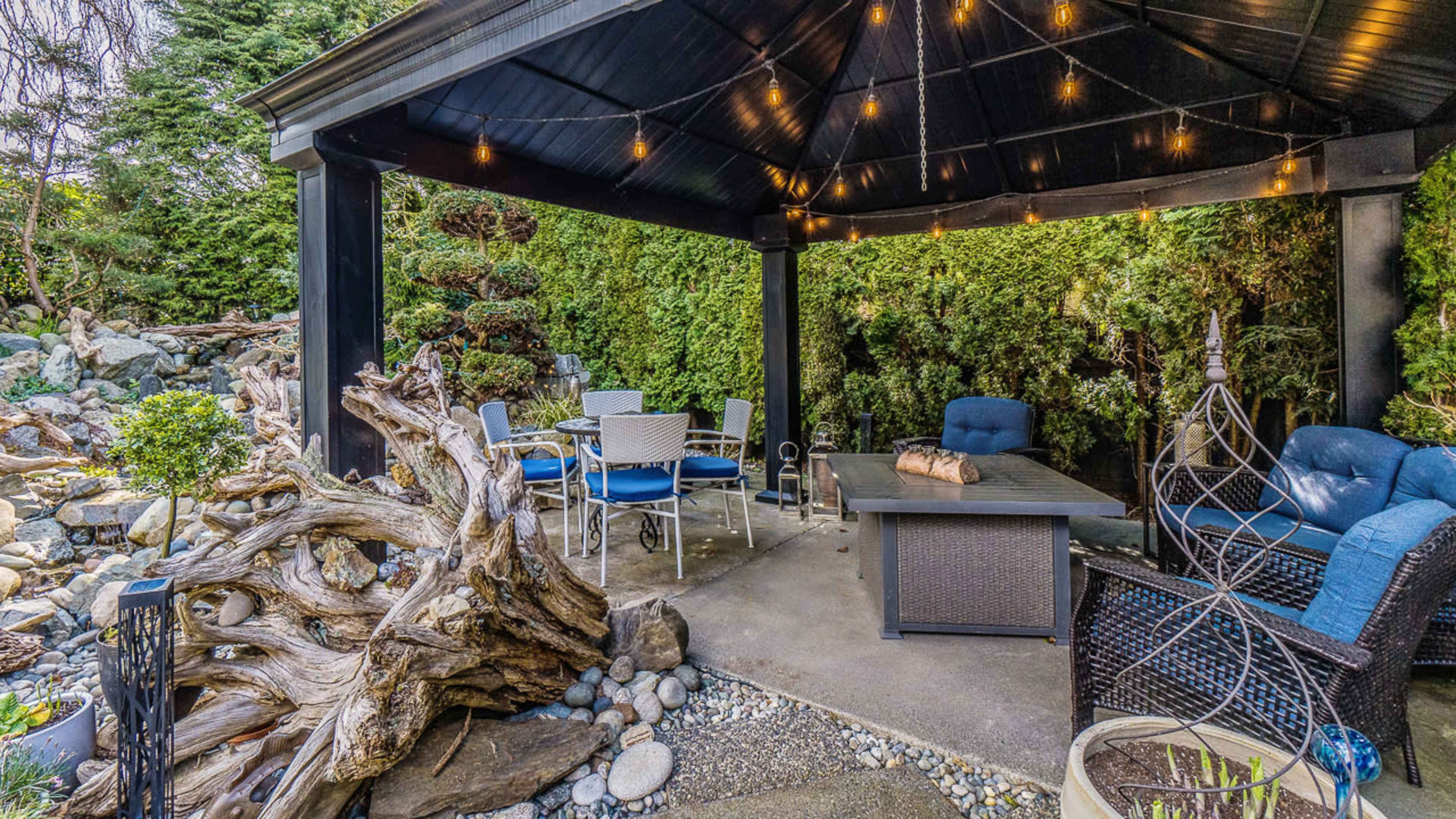 The image shows a covered outdoor seating area with a table and chairs, surrounded by lush greenery and decorative elements.