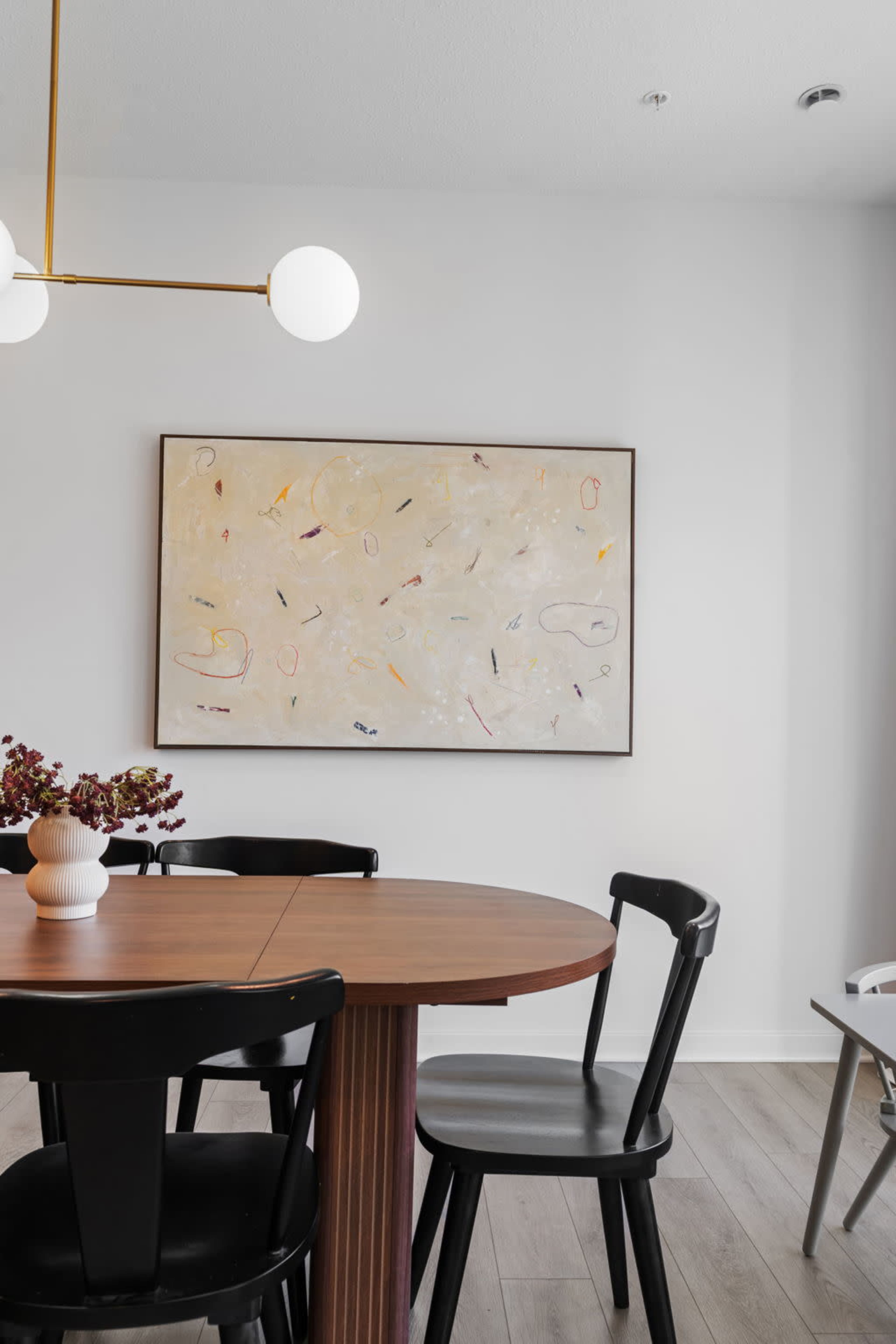 A wooden dining table surrounded by black chairs is positioned under a round light fixture, with a large abstract painting hanging on the wall.