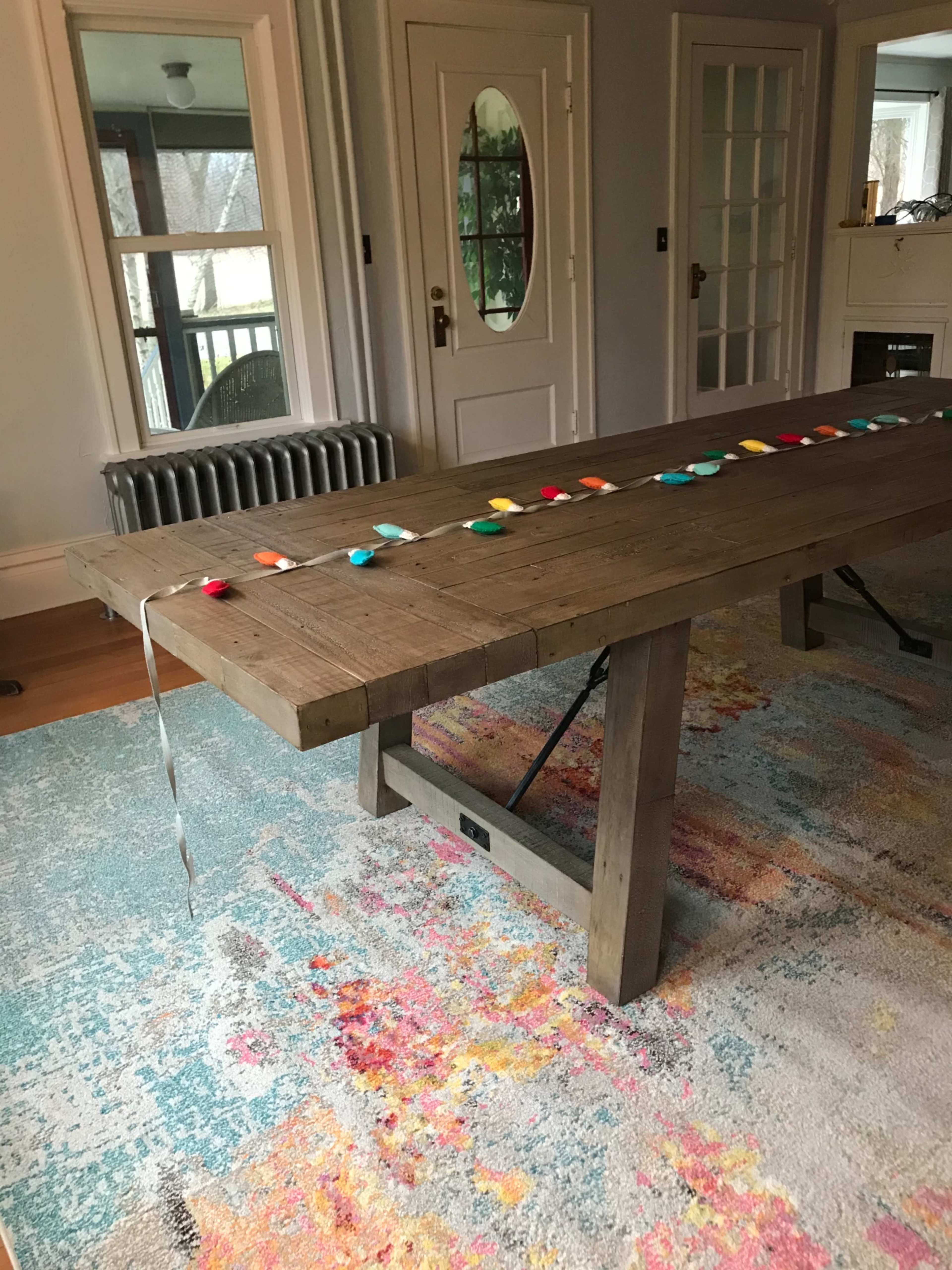 A wooden dining table is positioned on a colorful rug, with a string of multi-colored stones arranged along its center.