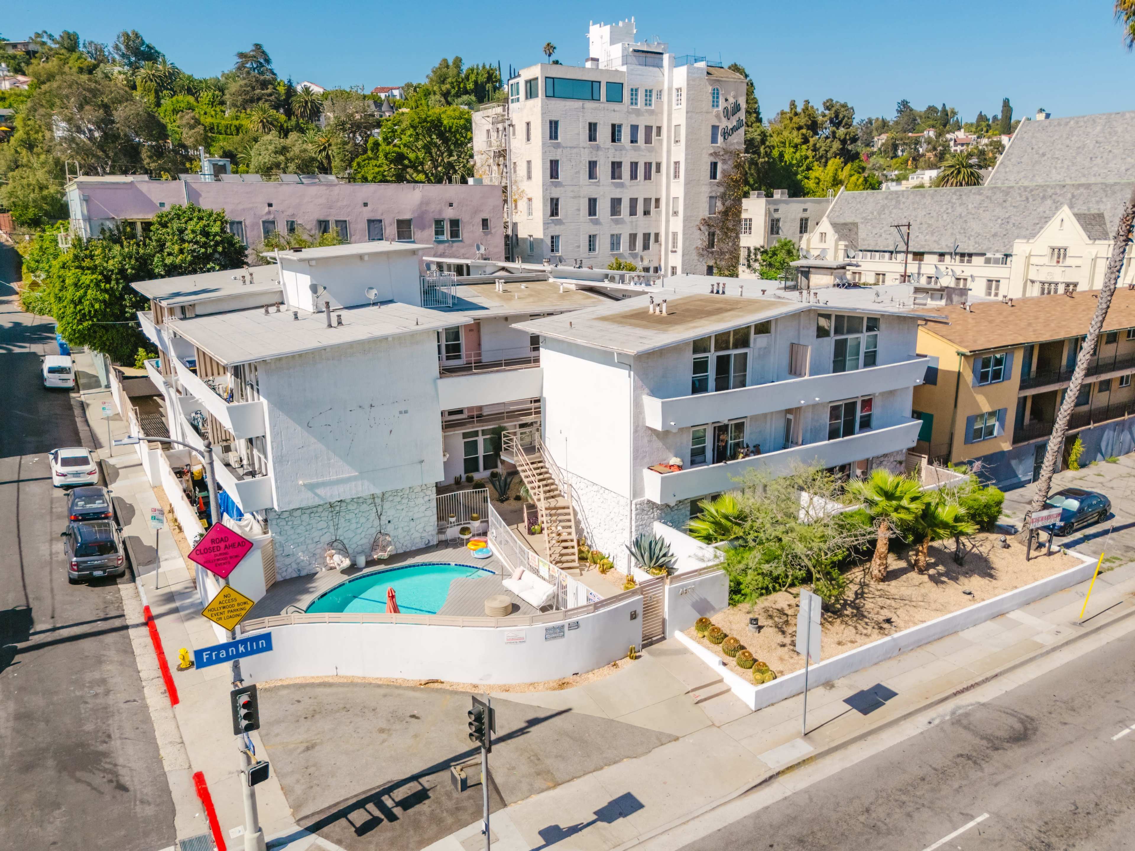 The image shows a mid-century style apartment building with a swimming pool, situated on a street corner surrounded by palm trees and other buildings.